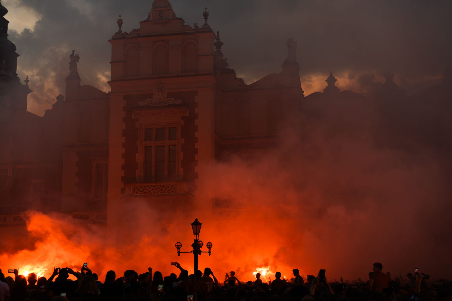 A silhouetted crowd gathers with burning orange flares in front of a historic building under a dark and cloudy sky.