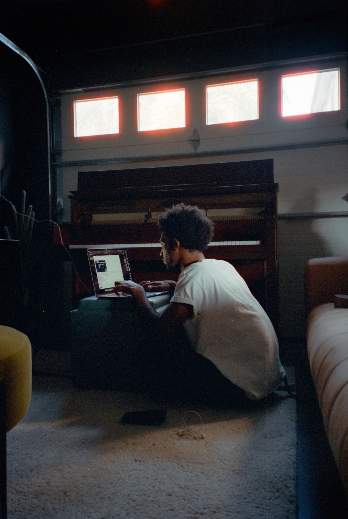 A young man sits on the carpeted floor in a dimly lit room, working on his laptop in front of a piano.