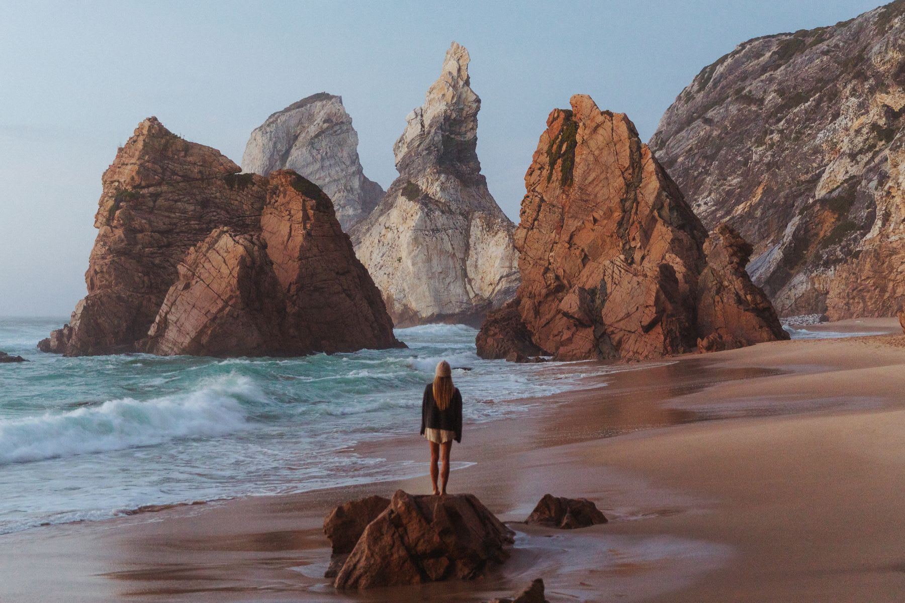 A woman stands on a rock at a dramatic and rugged beach, gazing out at massive sea stacks rising from the turquoise ocean.