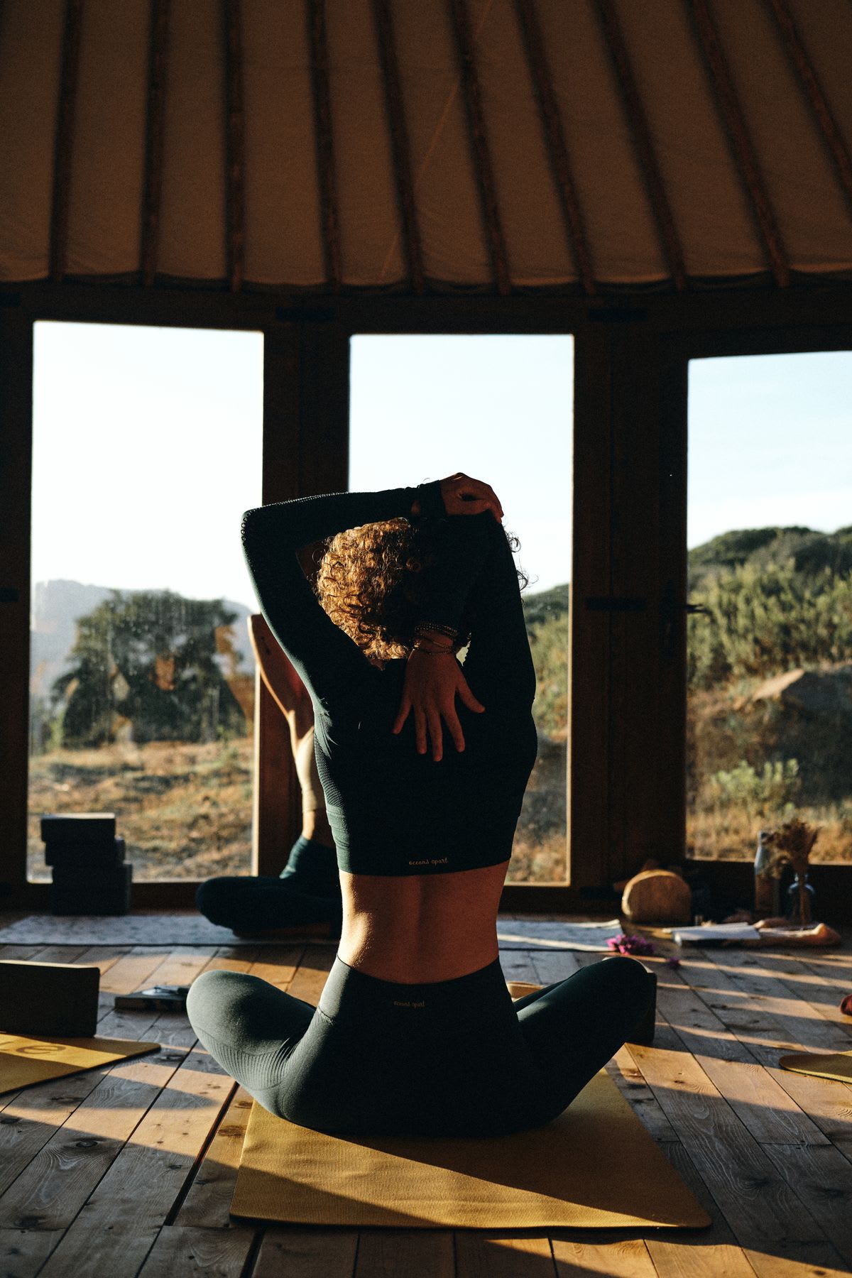 Woman practicing yoga in a sunlit environment, stretching in front of a large window that reveals a serene natural landscape.
