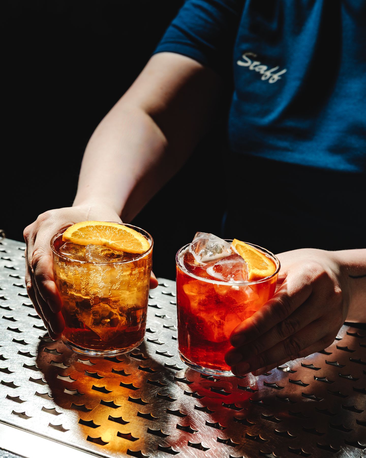 A bartender in a blue staff shirt presents two classic cocktails, a Negroni and an Old Fashioned, on a steel bar mat.