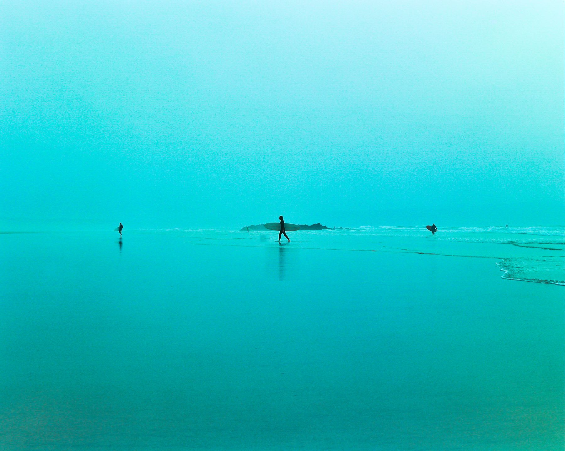 Silhouetted surfers with their boards walk along the shoreline of a tranquil turquoise ocean.