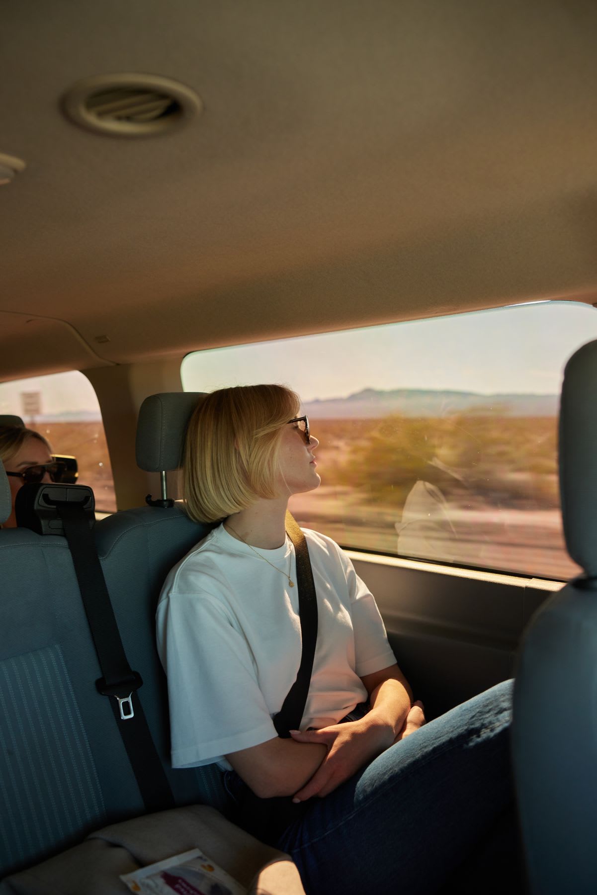 A woman wearing a white t-shirt sits strapped in with a seat belt, wearing sunglasses, looking through the car window during the day.