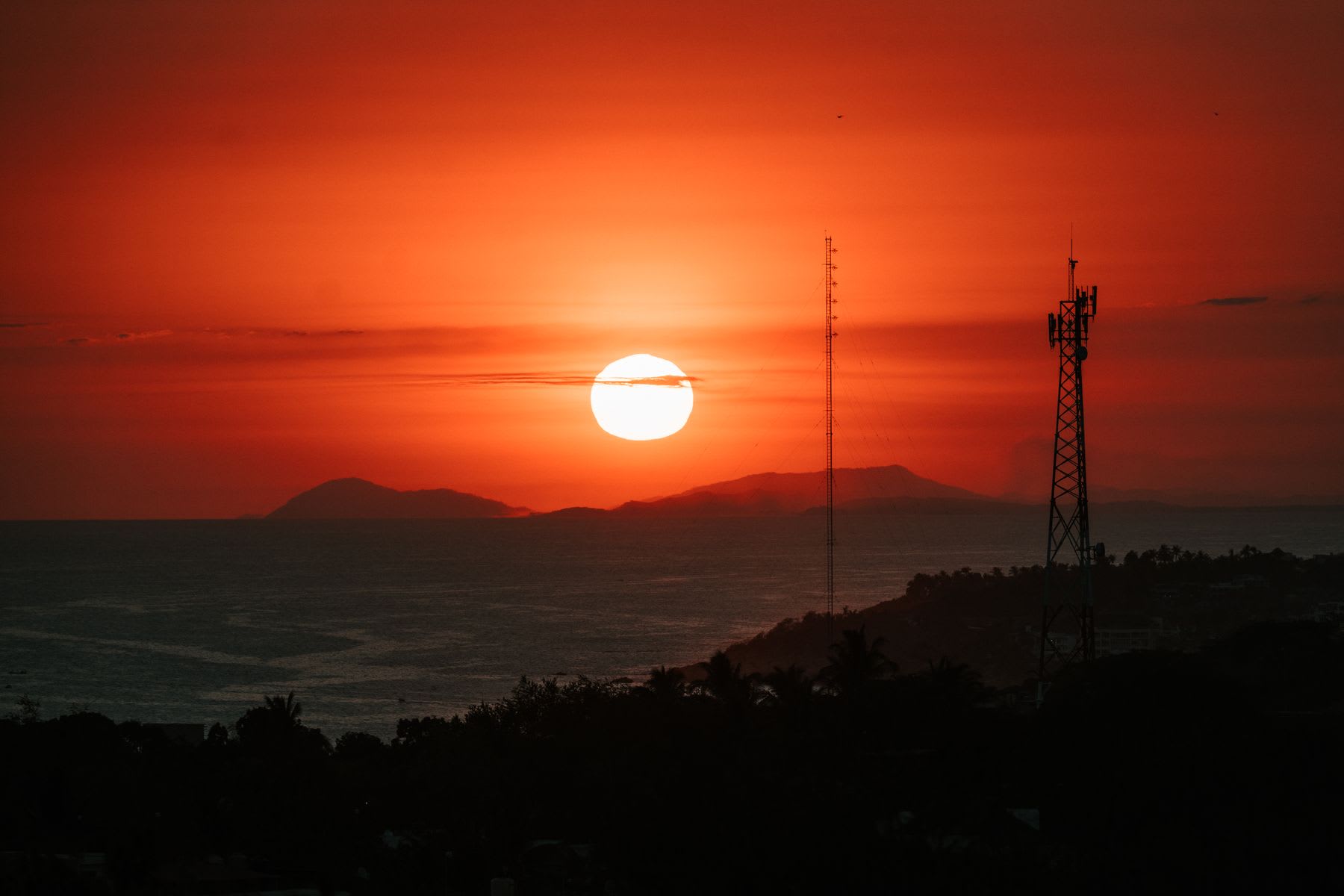 A vibrant orange sun sets over the ocean, silhouetting distant mountains and two communication towers on the shoreline.