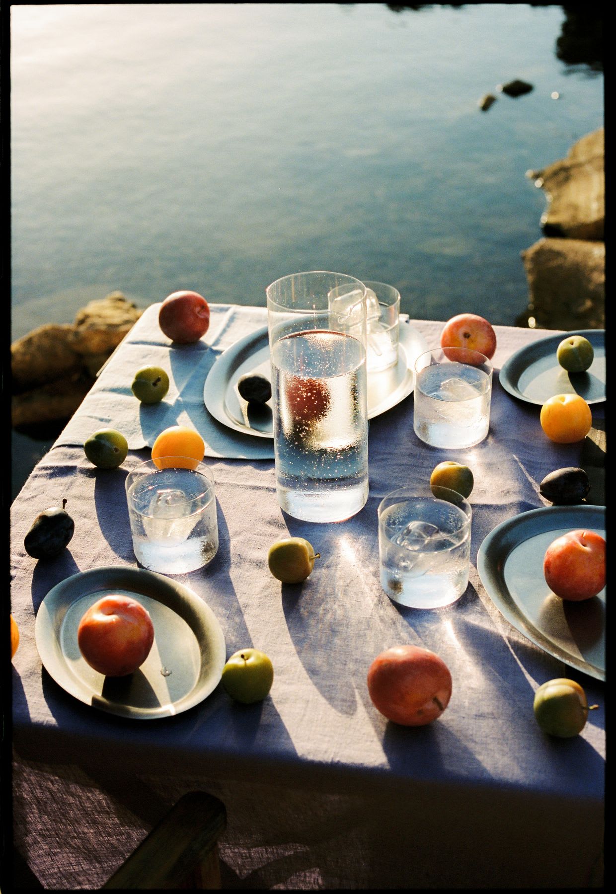 A sun-drenched table set with fresh fruit and glasses of sparkling water sits beside a calm body of water.