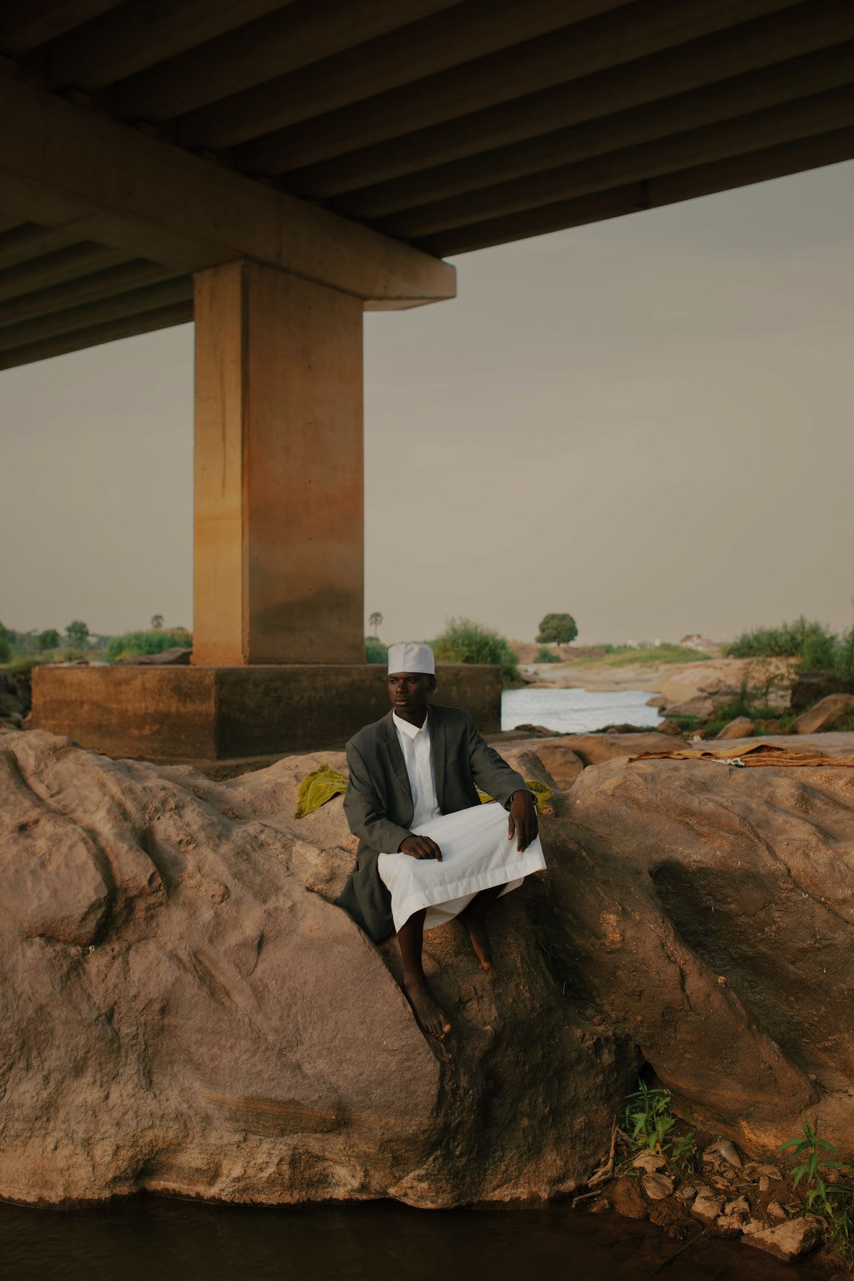 A man dressed in traditional clothing sits barefoot on a large rock formation under a concrete bridge.