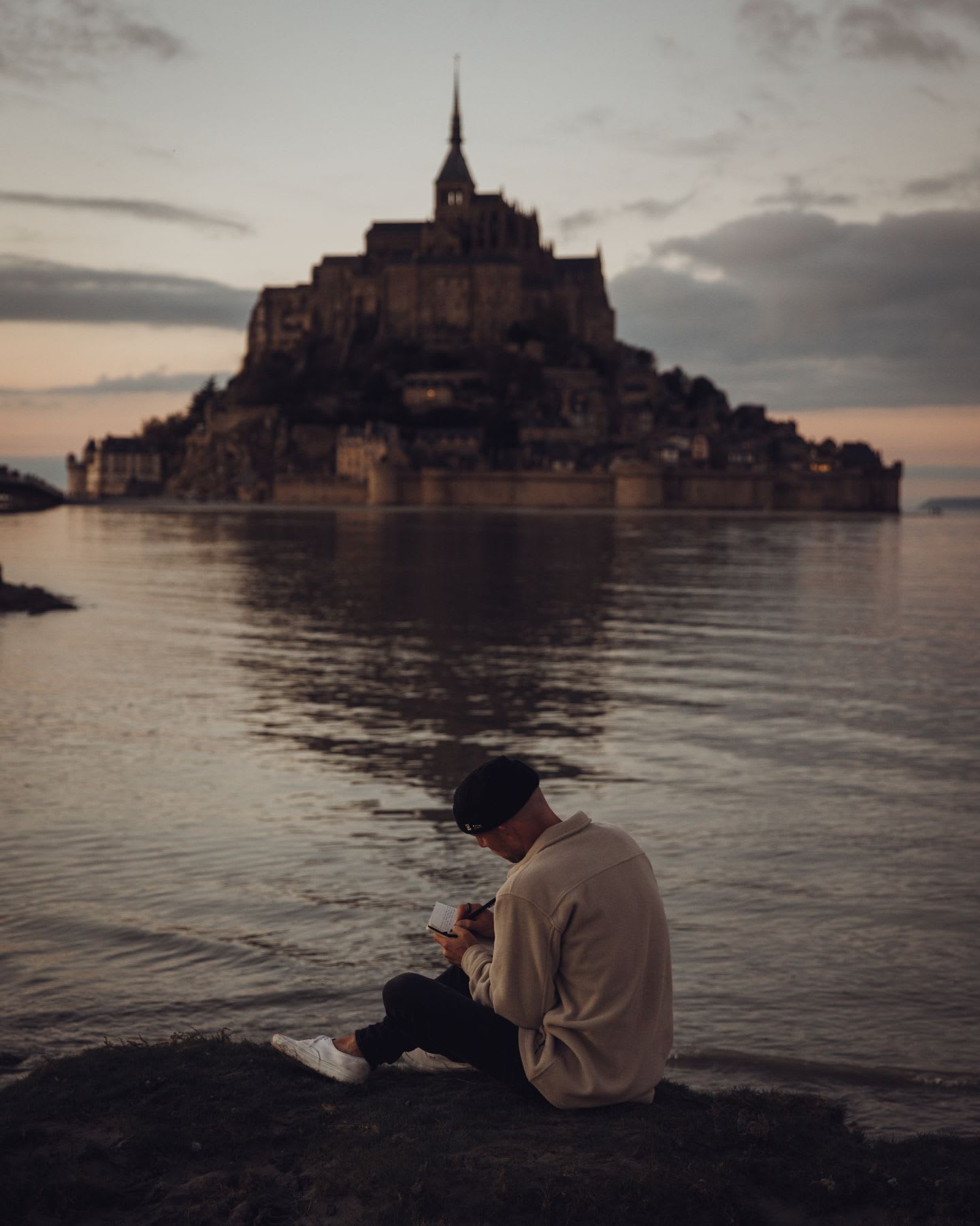 At dusk, a man in a cap and casual clothes sits on the shore writing in a notebook, with Mont Saint-Michel’s medieval abbey and fortifications silhouetted across the calm water behind him.