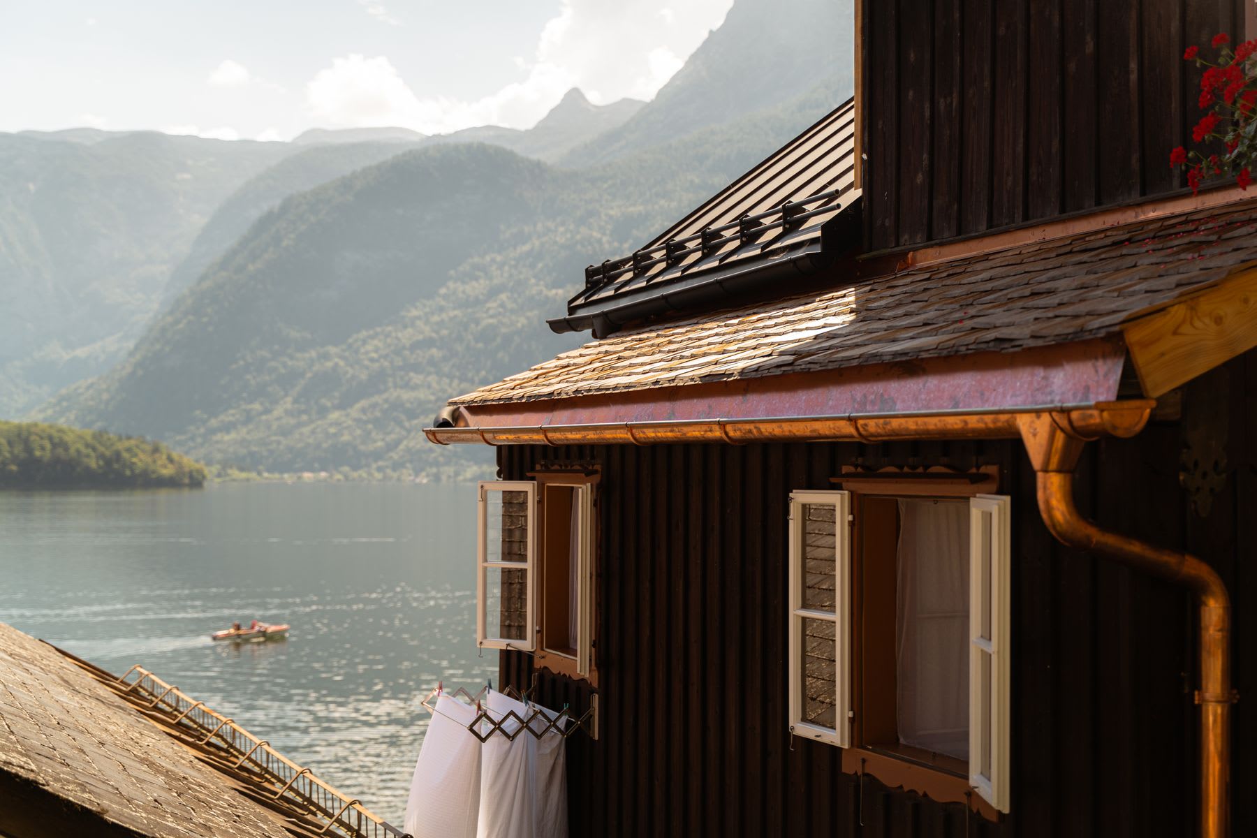 Wooden house with open windows and clothes drying in the sun next to a lake surrounded by mountains.