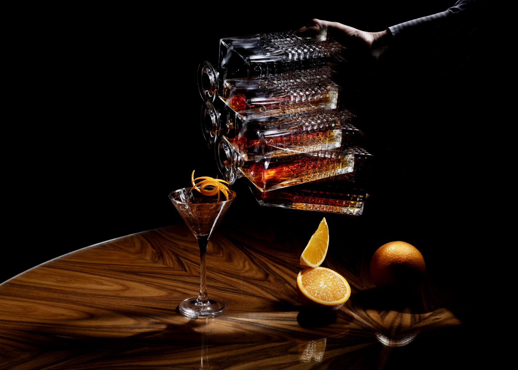 Several glasses with liquid flowing from one to the other on a wooden surface with an orange and its slice nearby.