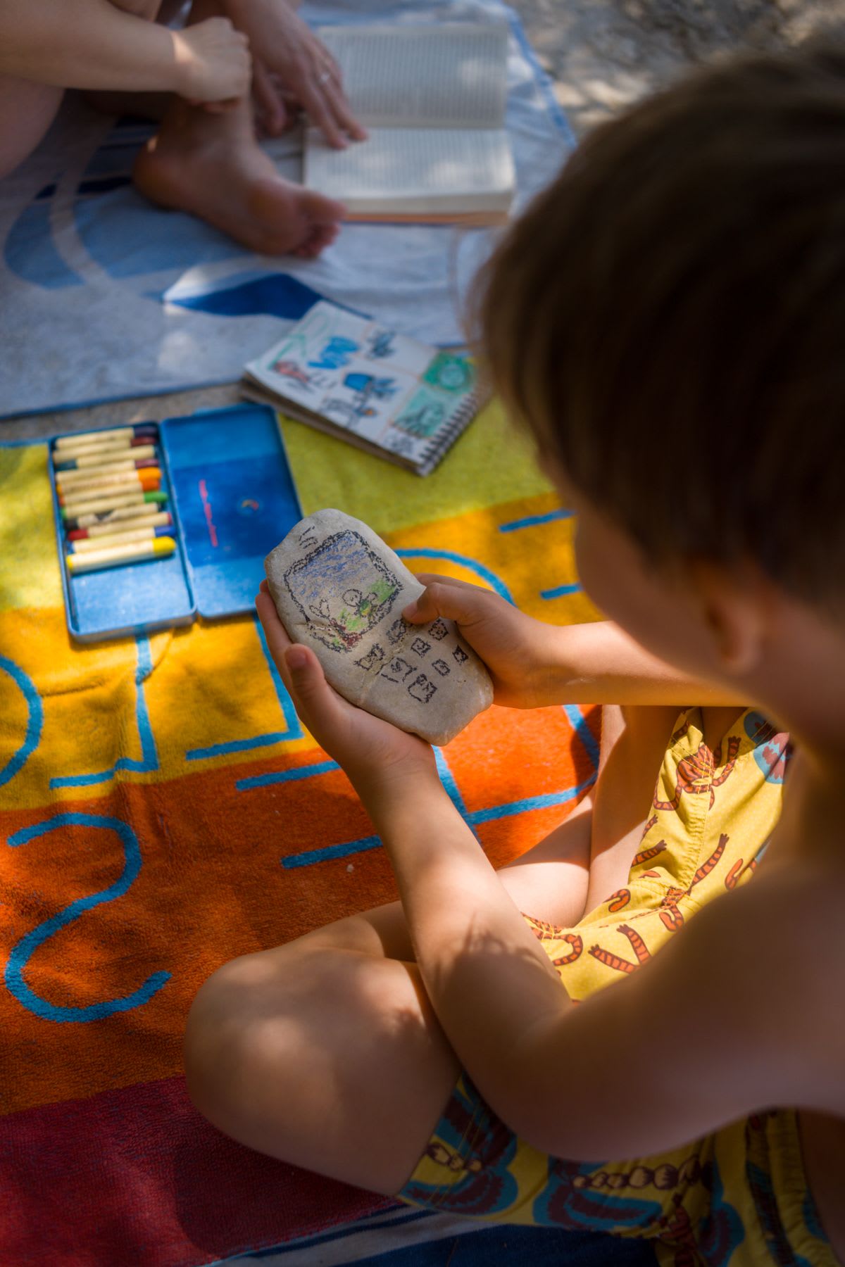 A child sits on a colorful towel, holding a painted rock that resembles a phone, with art supplies nearby.