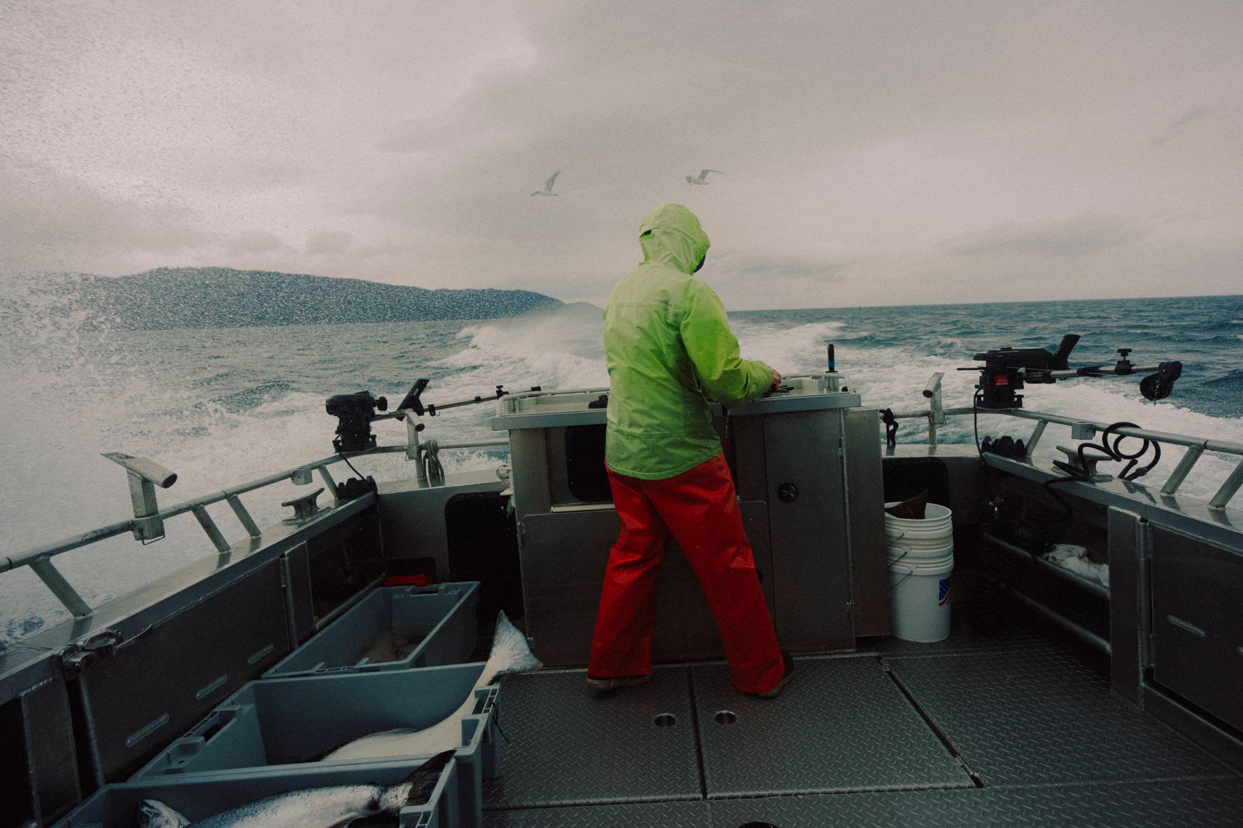 A person in a bright green rain jacket and red pants pilots a fishing boat through the choppy ocean with a fresh catch on deck.