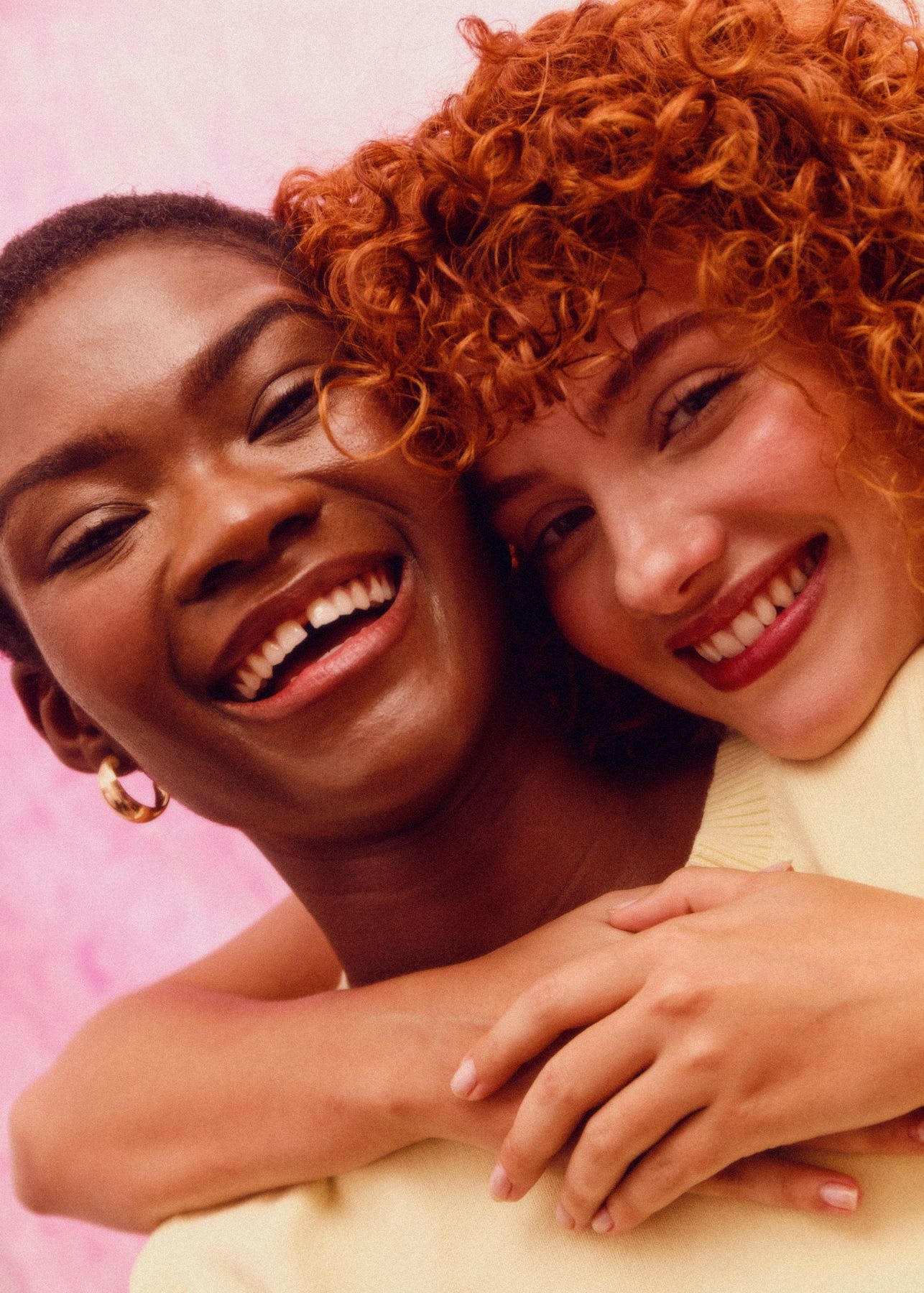 Two women are smiling and embracing closely. One woman has short, dark hair and wears hoop earrings, while the other has curly red hair.