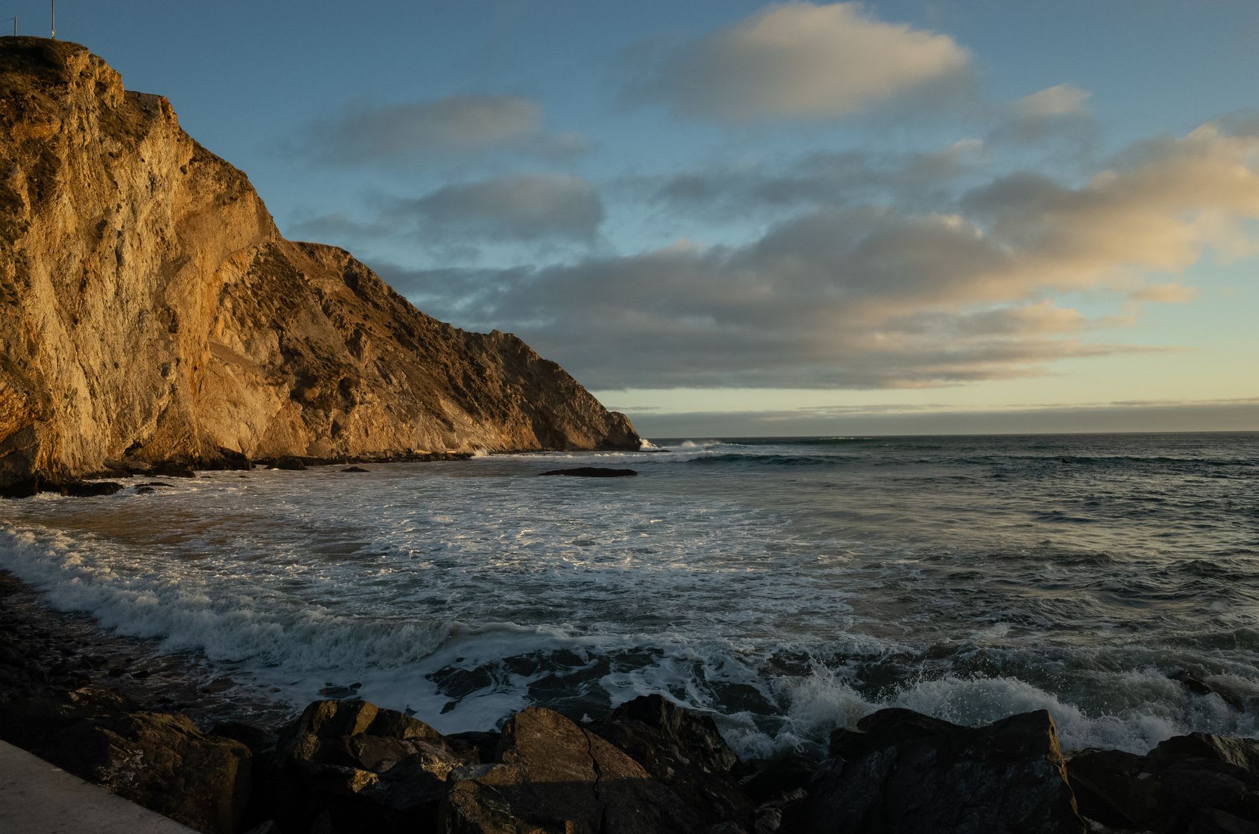 Golden light from the sun illuminates the rocky cliffs as waves crash on the shore of the ocean.