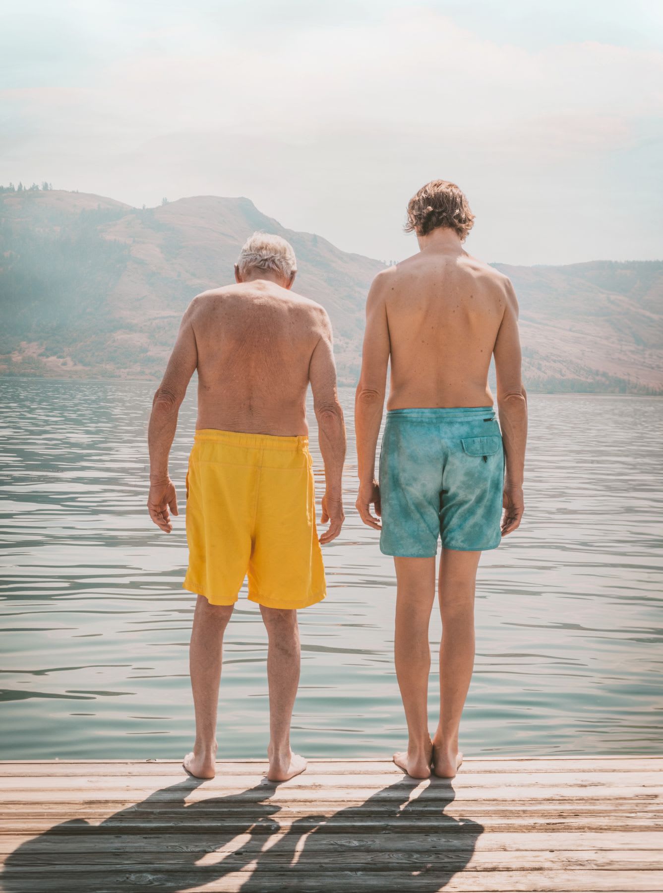 an elderly man and a younger man standing side by side on a wooden dock, facing a calm lake with distant hills in the background, both wearing swim trunks—one in yellow and the other in blue.