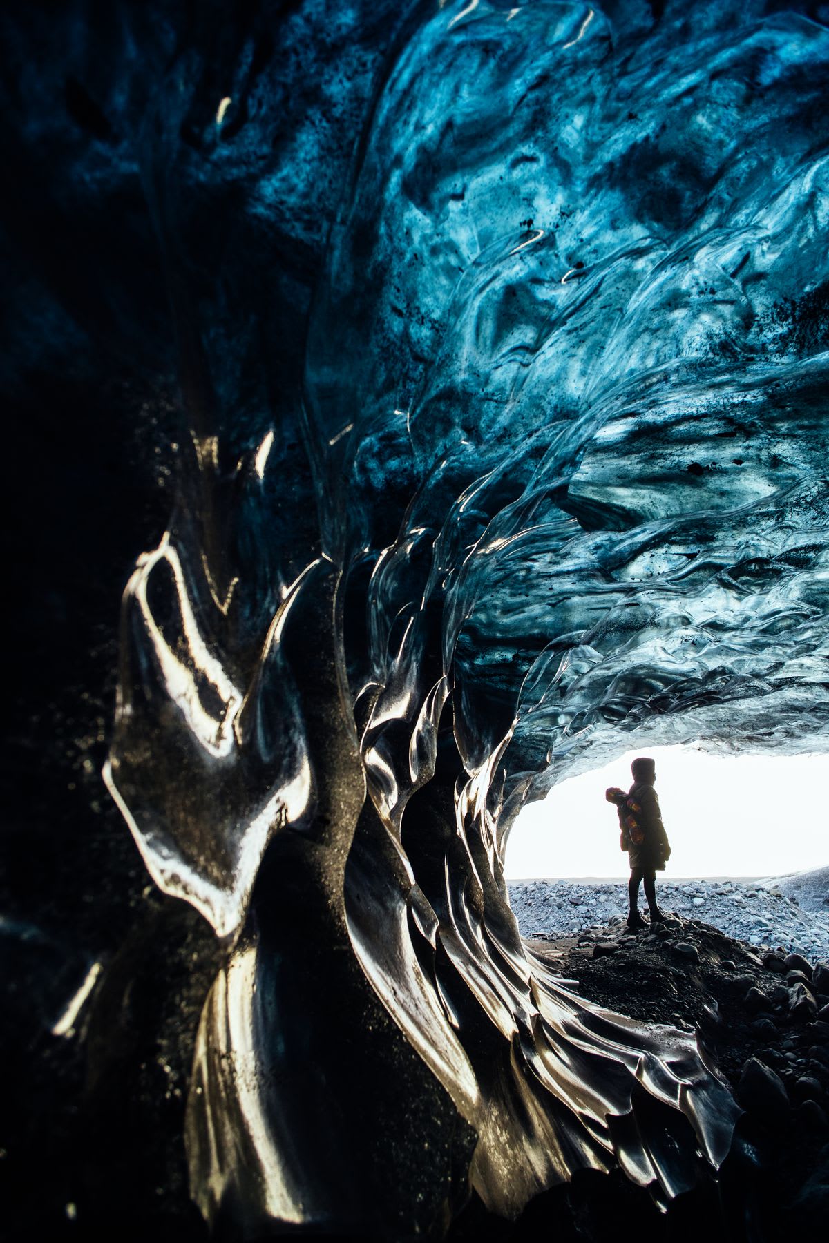 Person standing next to an ice cave with blue translucent walls and frozen wave textures.