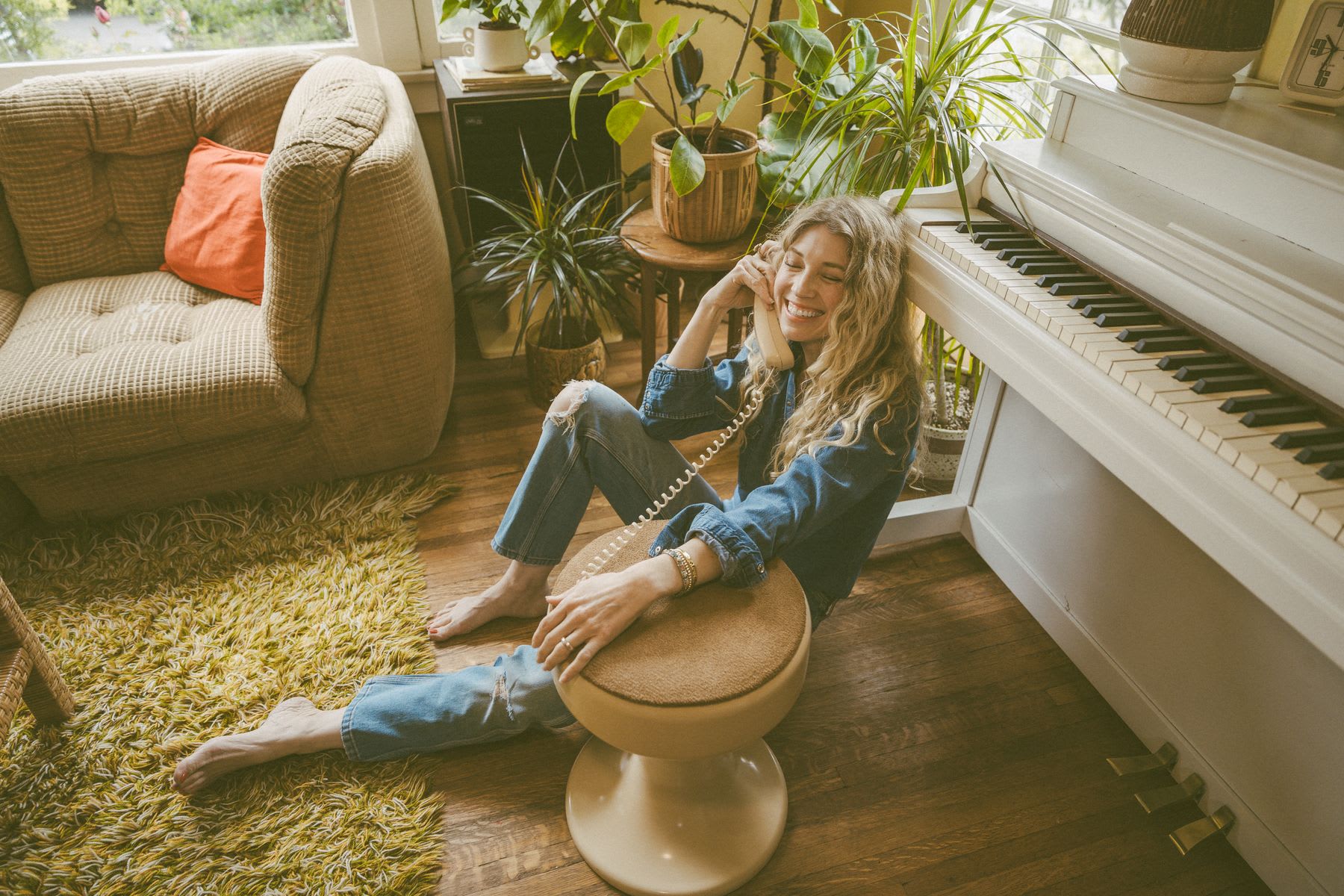 A smiling woman with long, blonde hair, wearing a denim shirt and ripped jeans, sits barefoot on a wooden floor in a cozy, plant-filled room, talking on a rotary phone, with a white piano and a textured armchair in the background.