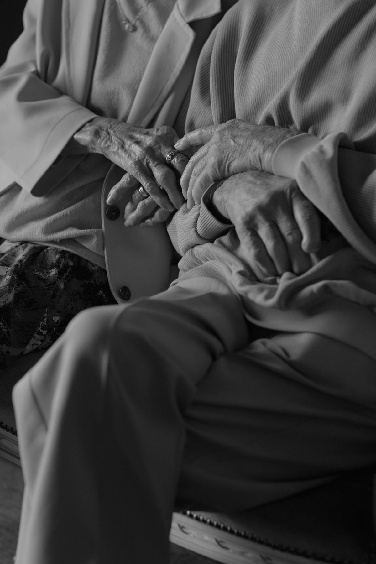 A close-up, black and white photograph captures the intertwined hands of an elderly couple, conveying a sense of comfort and intimacy.