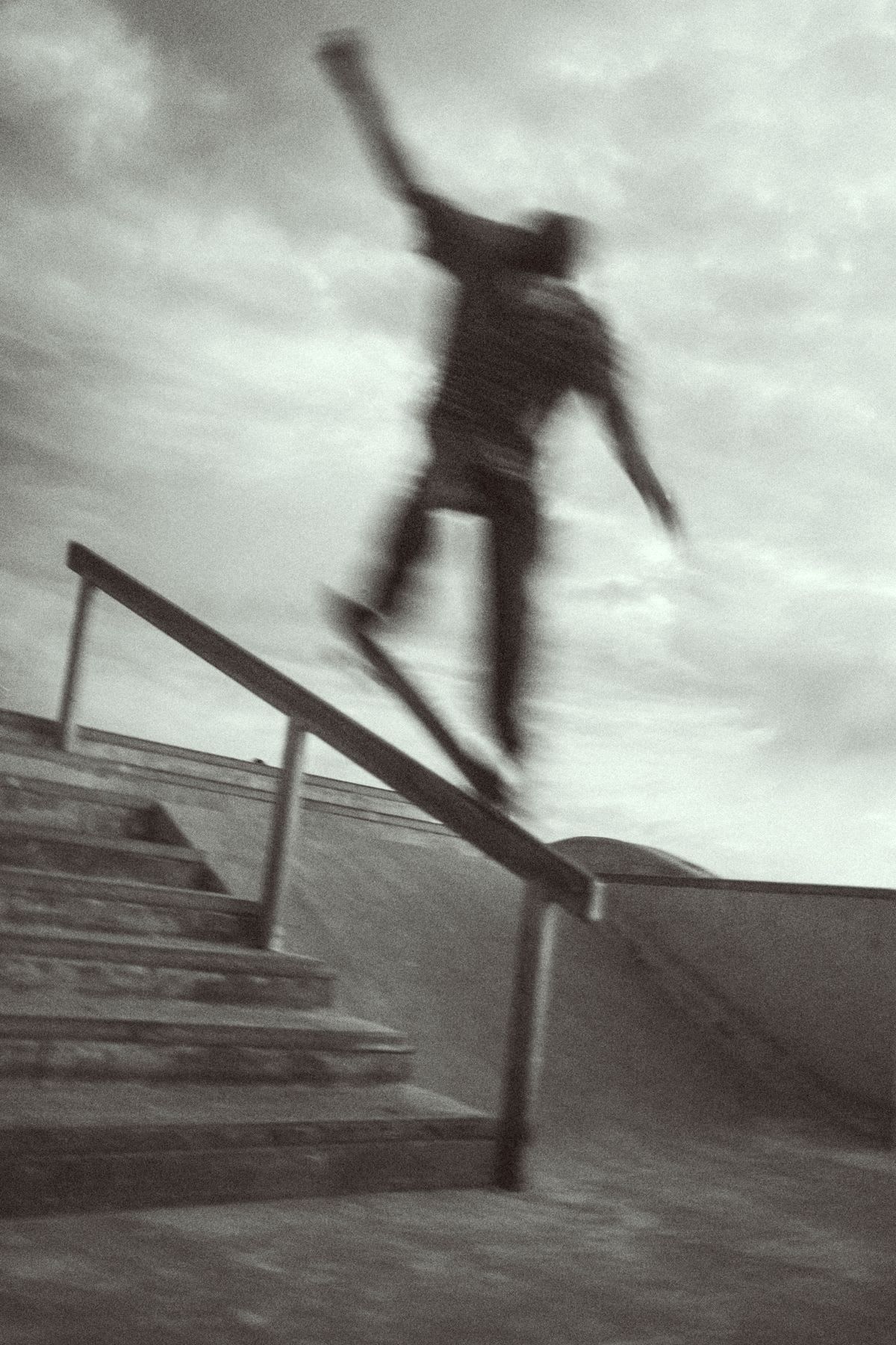 A motion-blurred, black and white photograph captures a skateboarder grinding down a handrail next to a concrete staircase.