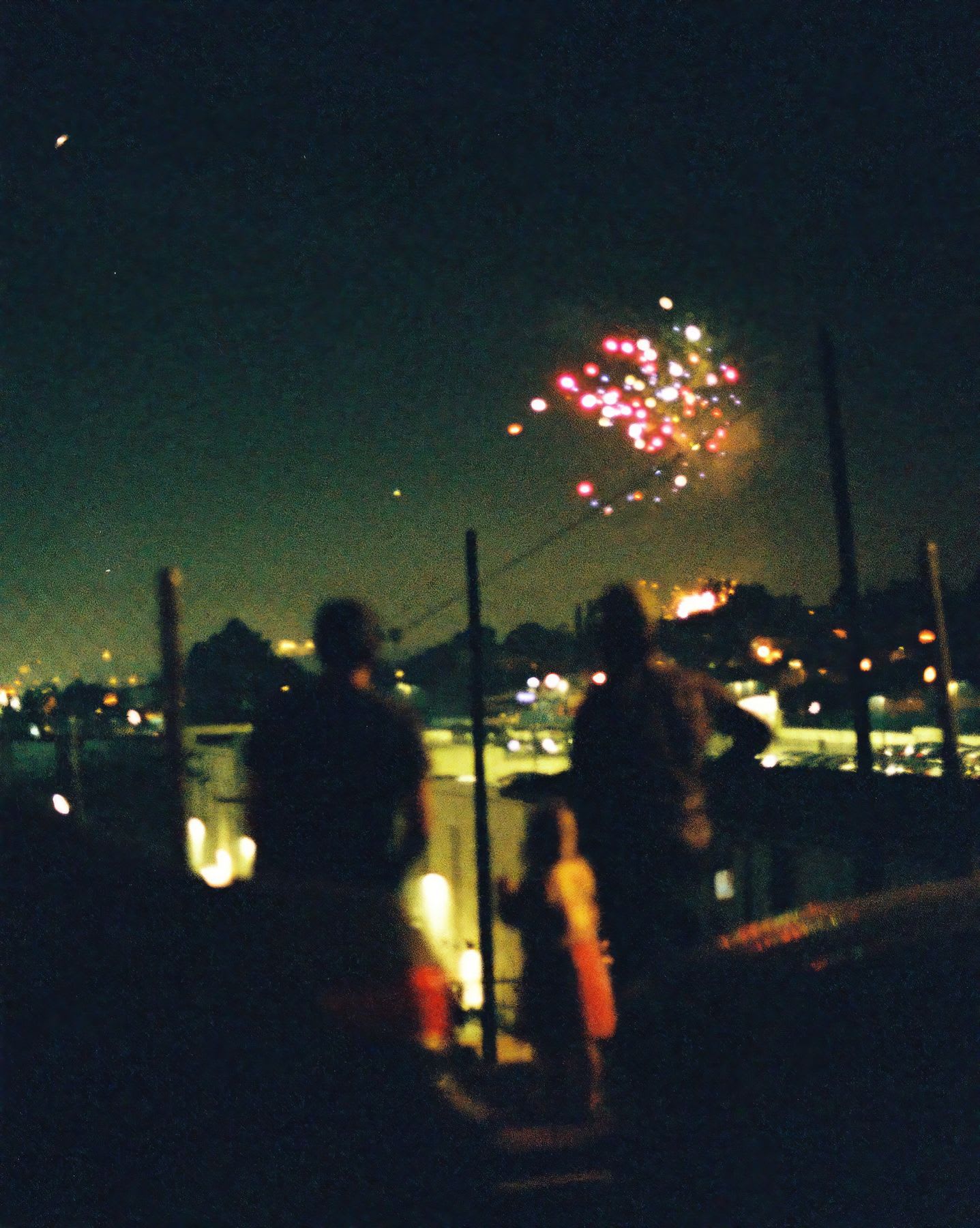 Silhouettes of people watch as colorful fireworks explode in the dark night sky over a city.