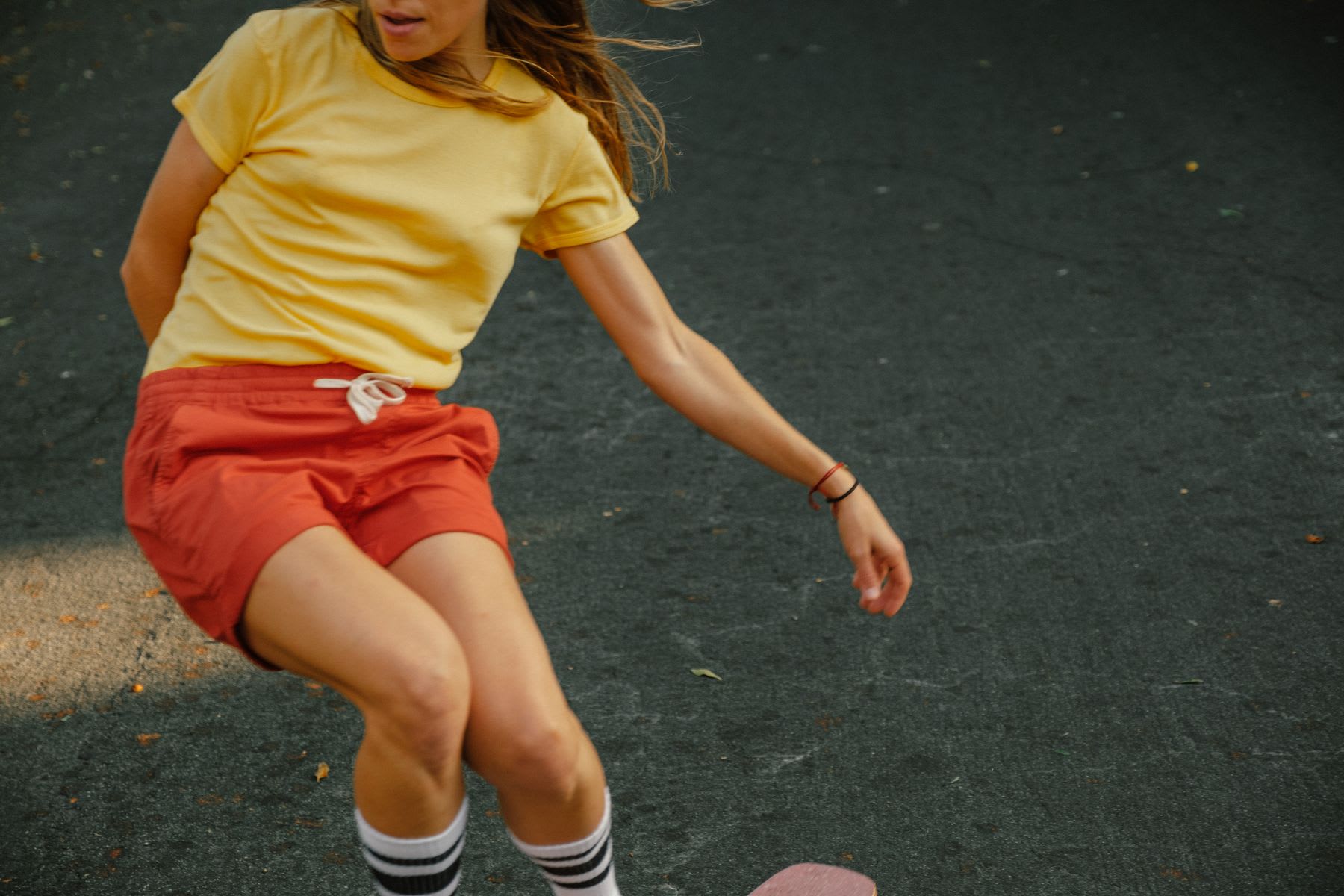 A woman wearing a yellow t-shirt, red shorts, and high-top socks rides a skateboard on an asphalt surface.