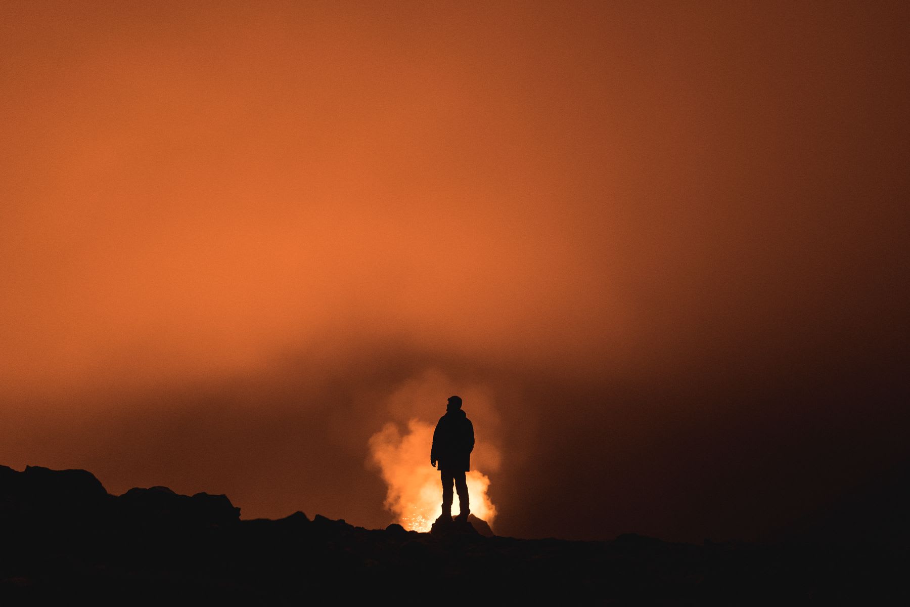 A man stands on a mountain at night with smoke rising into the red sky