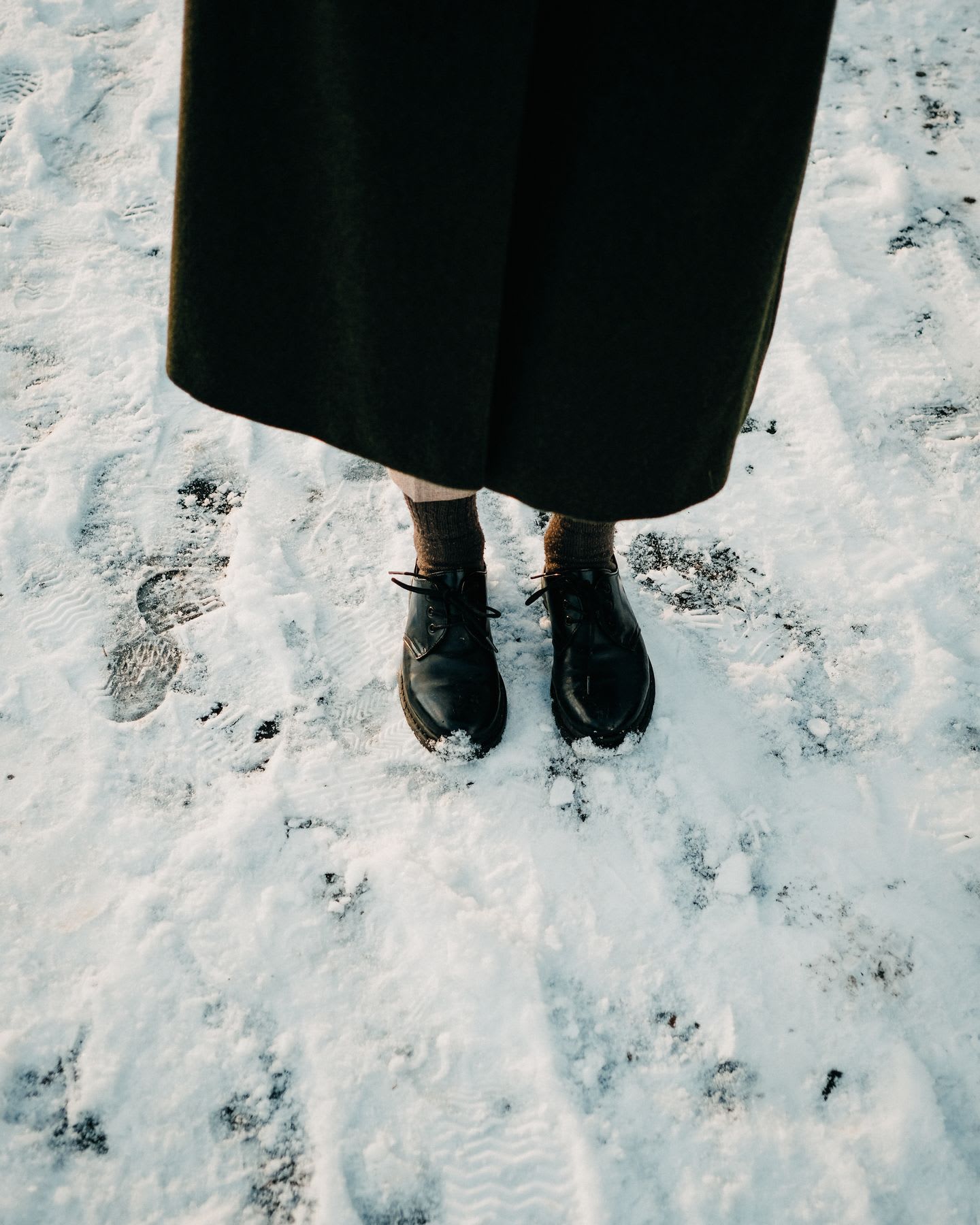 A high-angle view captures a person wearing black leather shoes and a long dark coat standing in the snow.
