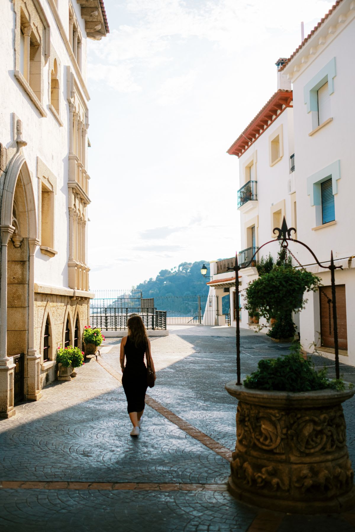 A woman in a black dress leisurely walking down a picturesque town street, with vibrant flowers blooming along the sidewalk.