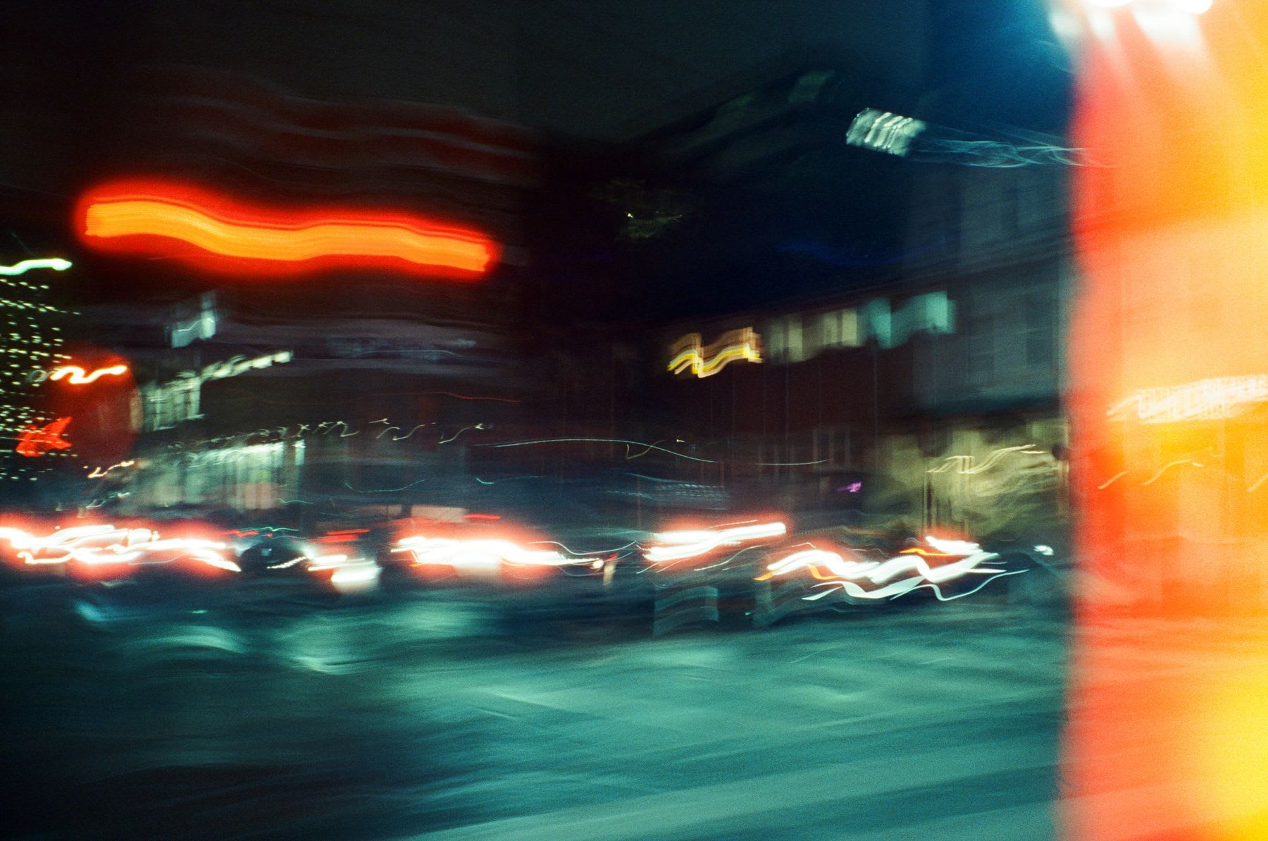 Blurred long exposure of a nighttime city street, with streaks of red, yellow, and white lights from cars and buildings.