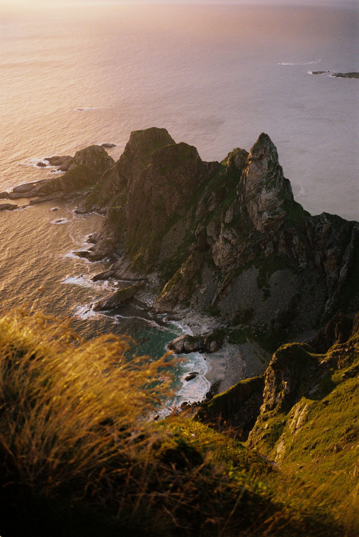 Rocky cliffs covered in vegetation slope down to the calm sea, illuminated by the golden light of the sunset.