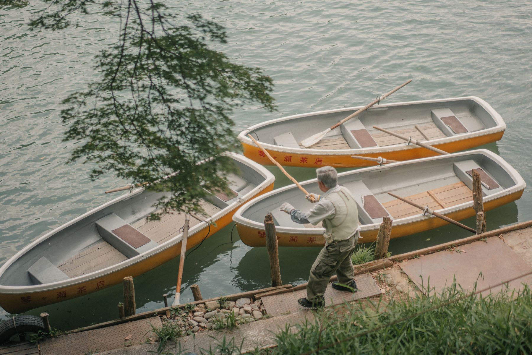 A man stands on a pier, holding an oar, next to three wooden boats moored to the shore of a lake.