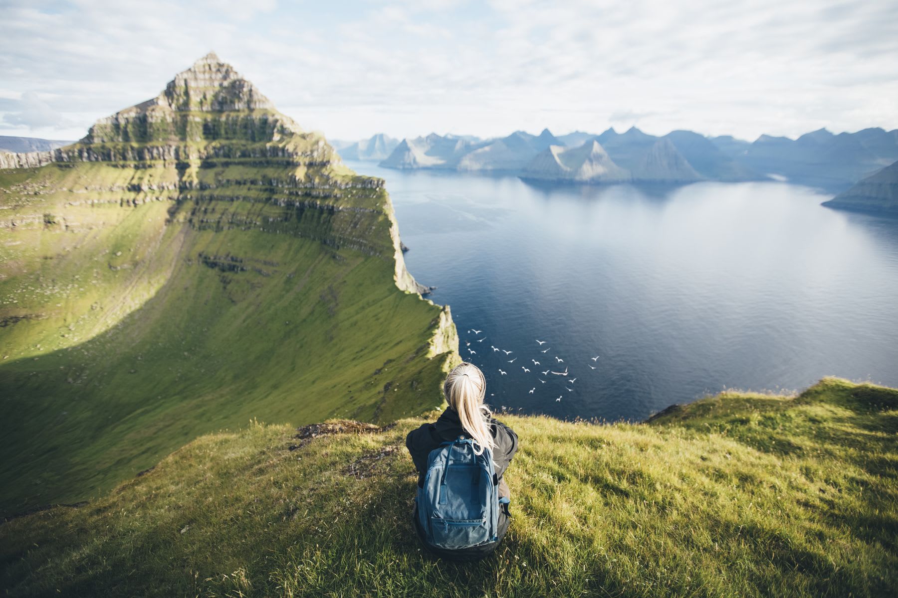 Blonde hiker sits on ground overlooking ridge and lake on cloudy day