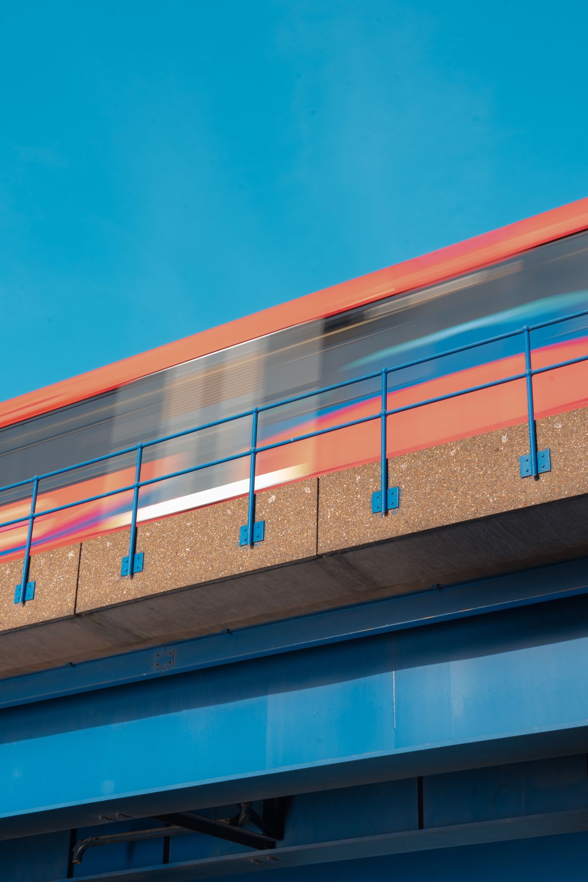 An orange train in motion blurs as it speeds across a blue overpass against a clear sky.