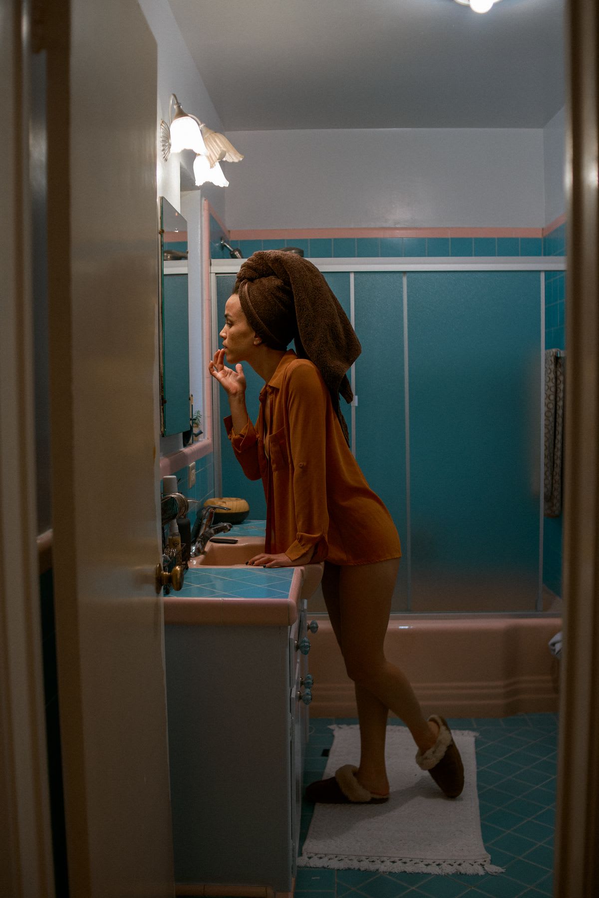 A woman with a towel wrapped around her head leans over the sink to apply makeup in a vintage-style bathroom.