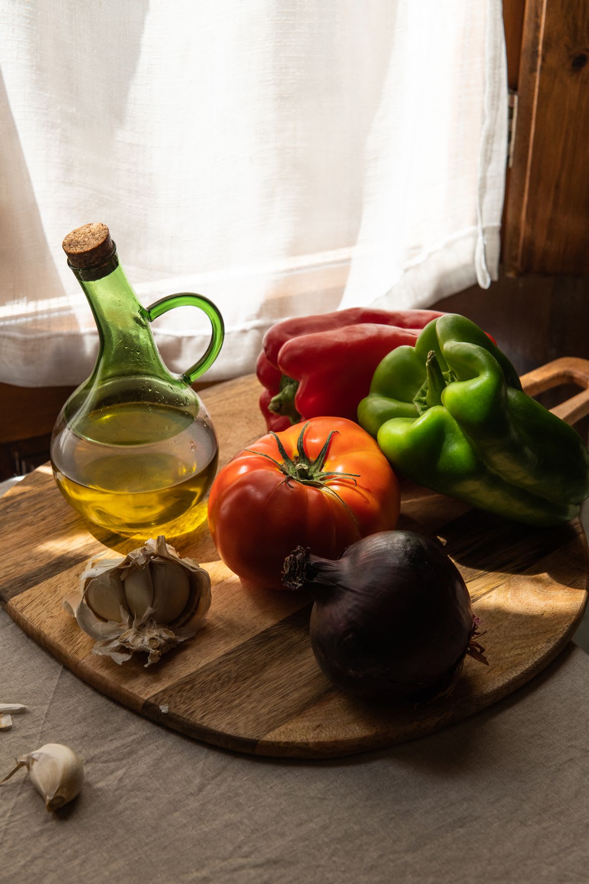 Bottle of olive oil with cork lid next to red and green peppers, ripe tomatoes, red onion and garlic cloves on a wooden board.
