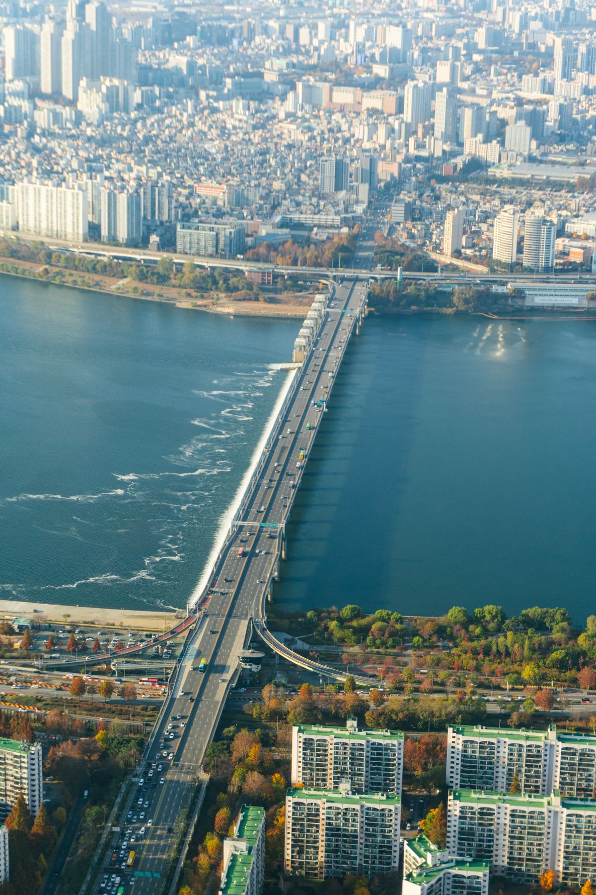 An aerial view shows a sprawling metropolis with a long highway bridge crossing a wide river under the bright daylight.