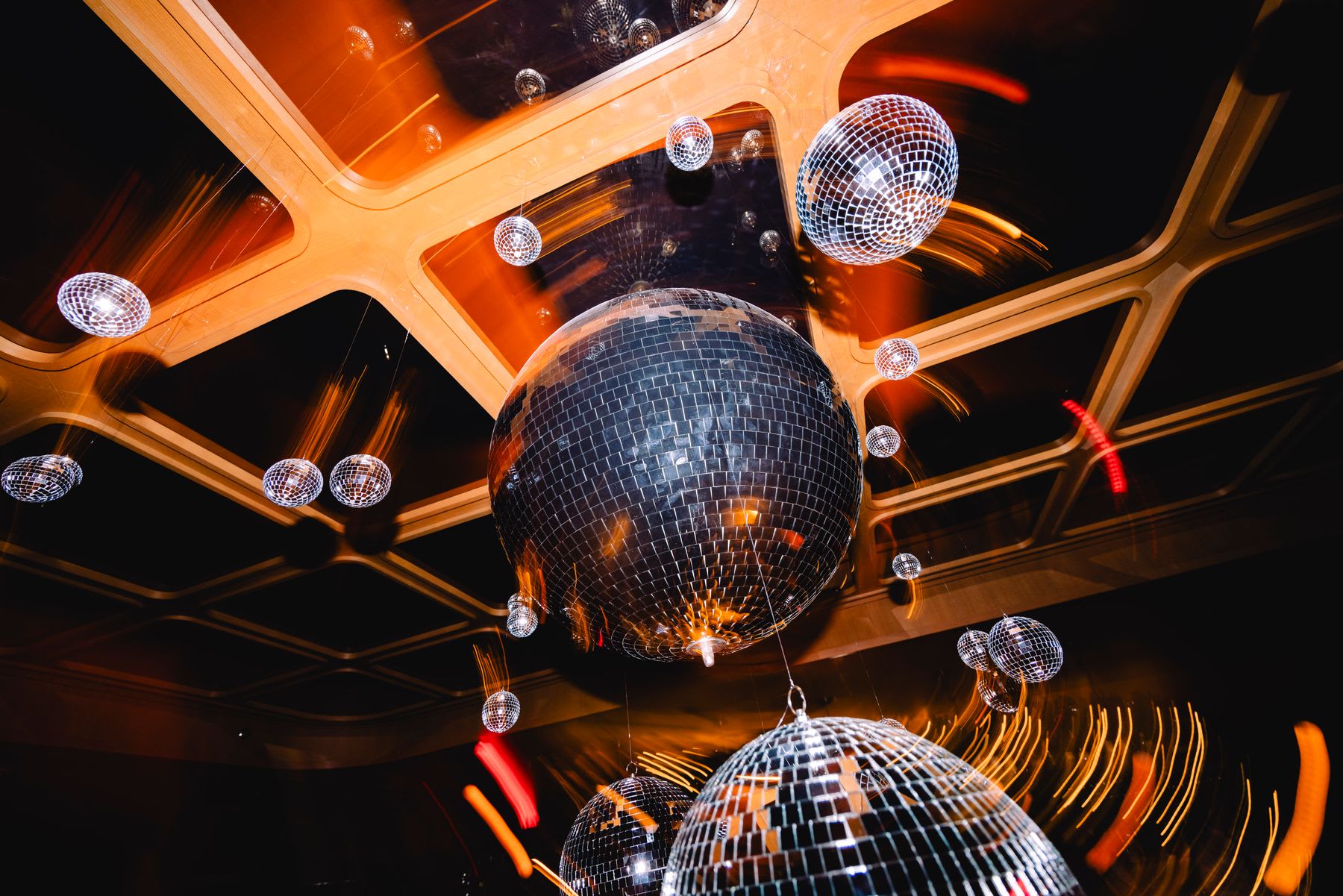 A vibrant display of various sized disco balls hangs from a modern, geometrically designed ceiling, bathed in warm, glowing light.
