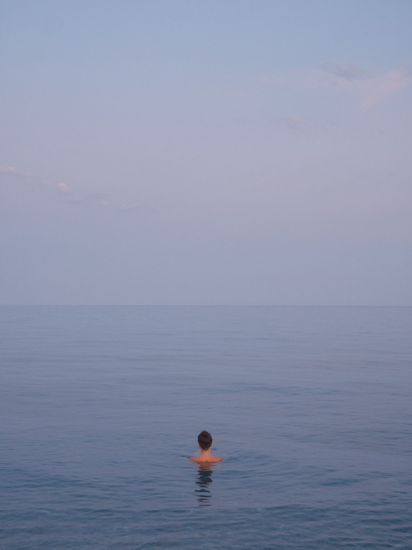 A lone person seen from the back swims in the vast, calm sea, with the water blending into the pale sky at the horizon.