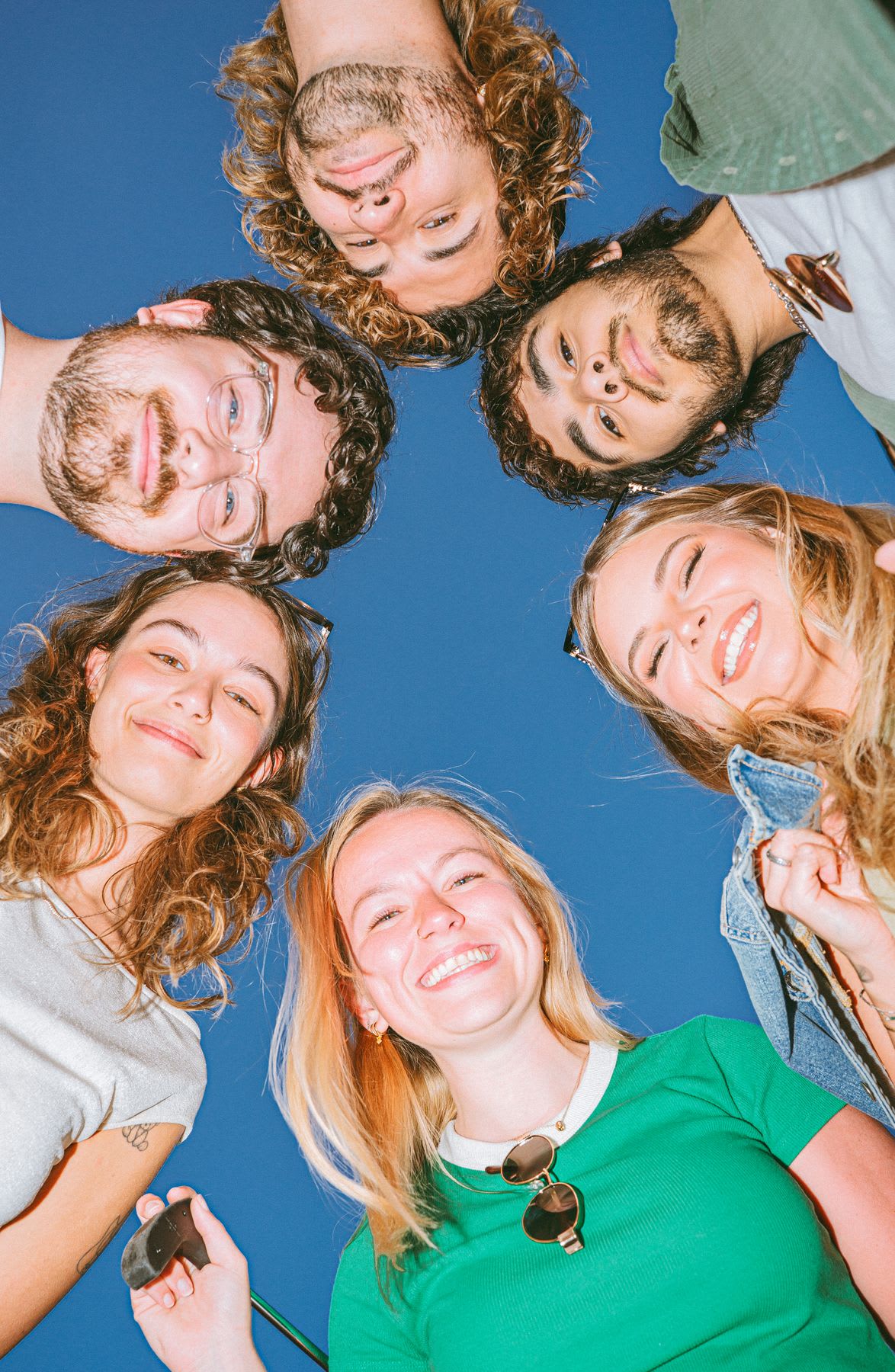 A low-angle view of a diverse group of six happy young friends huddled in a circle, smiling down at the camera against a clear blue sky.
