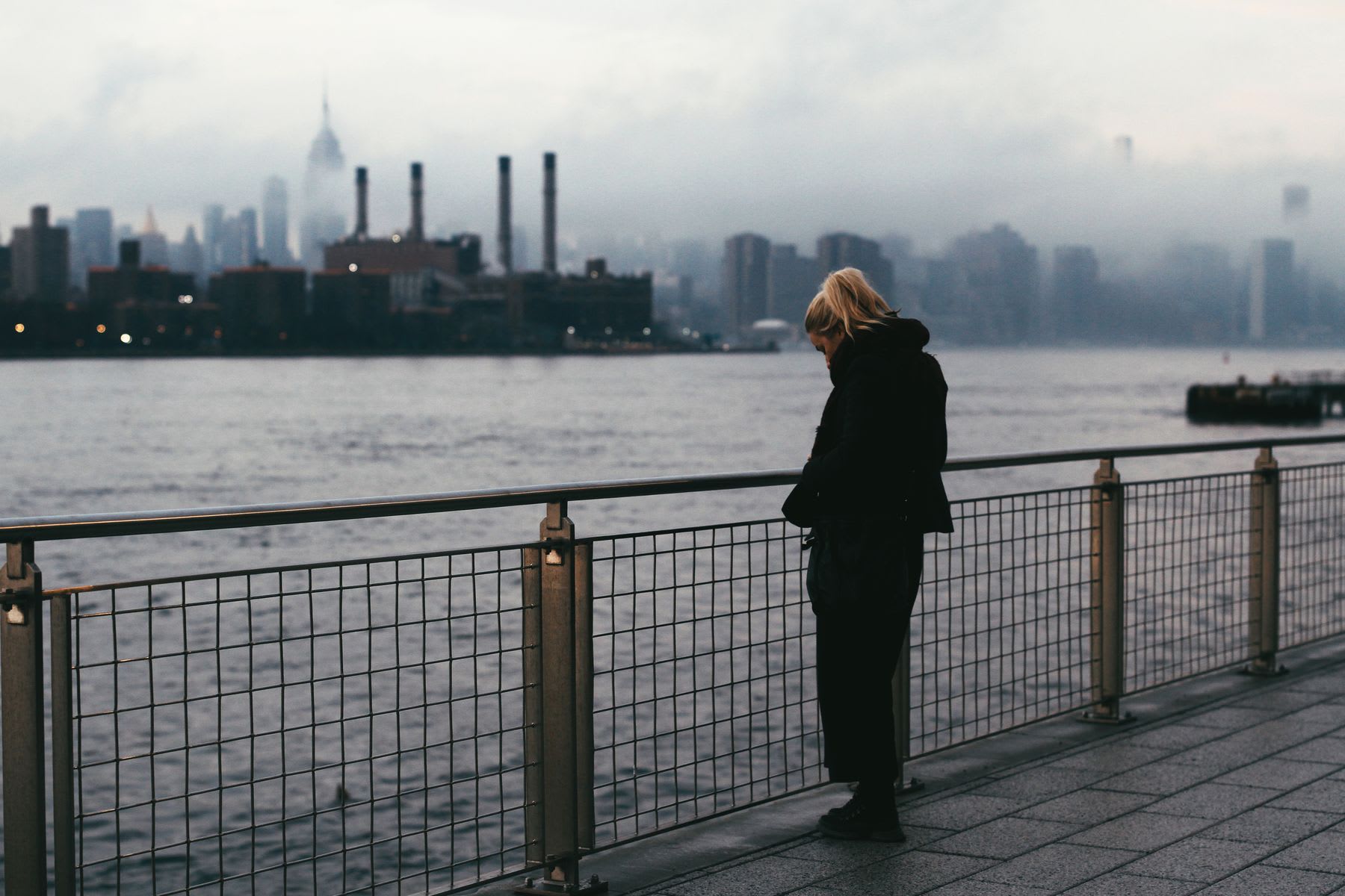 A women is standing on a pier looking into the water wearing warm clothes.