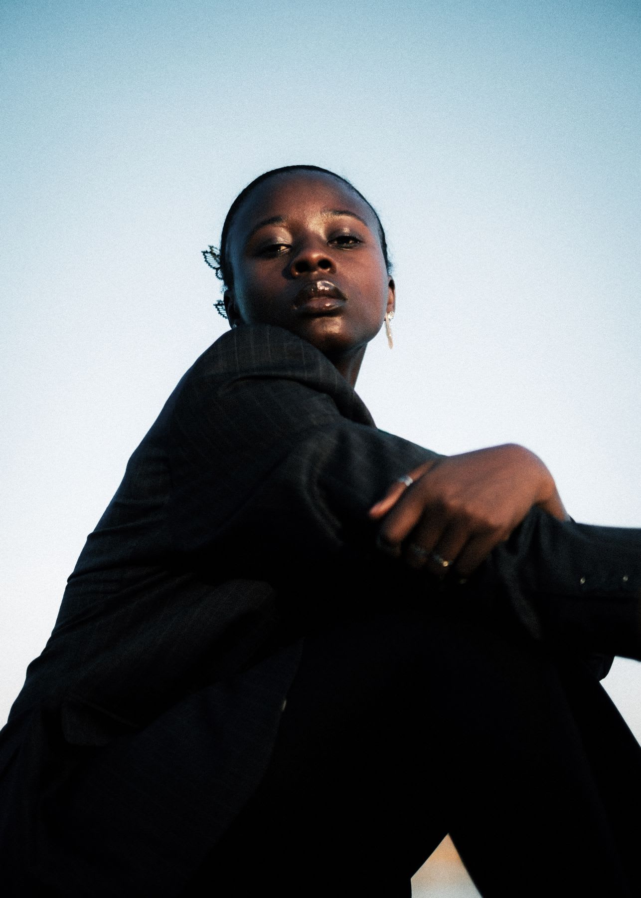 A low-angle portrait of a confident young Black woman in a dark pinstripe blazer, looking directly at the camera while sitting against a clear sky.
