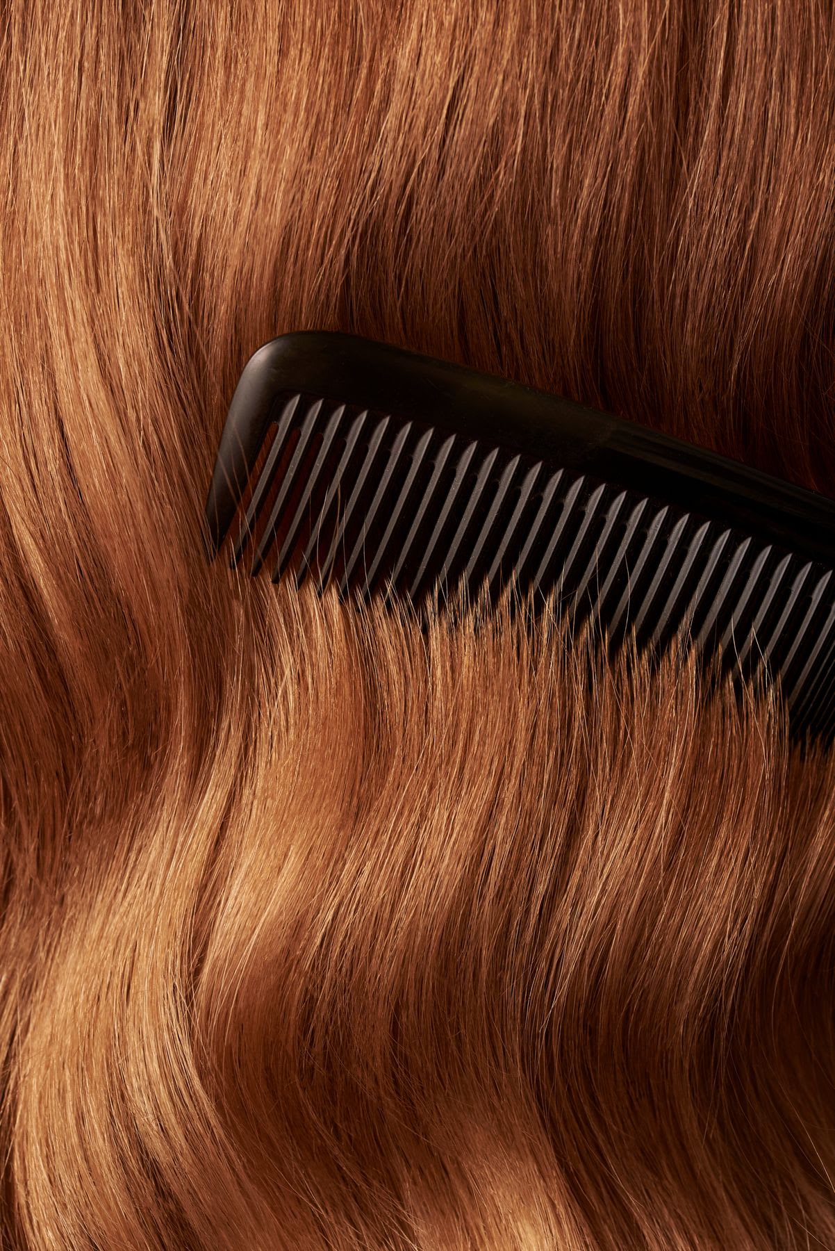 A close-up of a black comb running through thick and wavy light brown hair.
