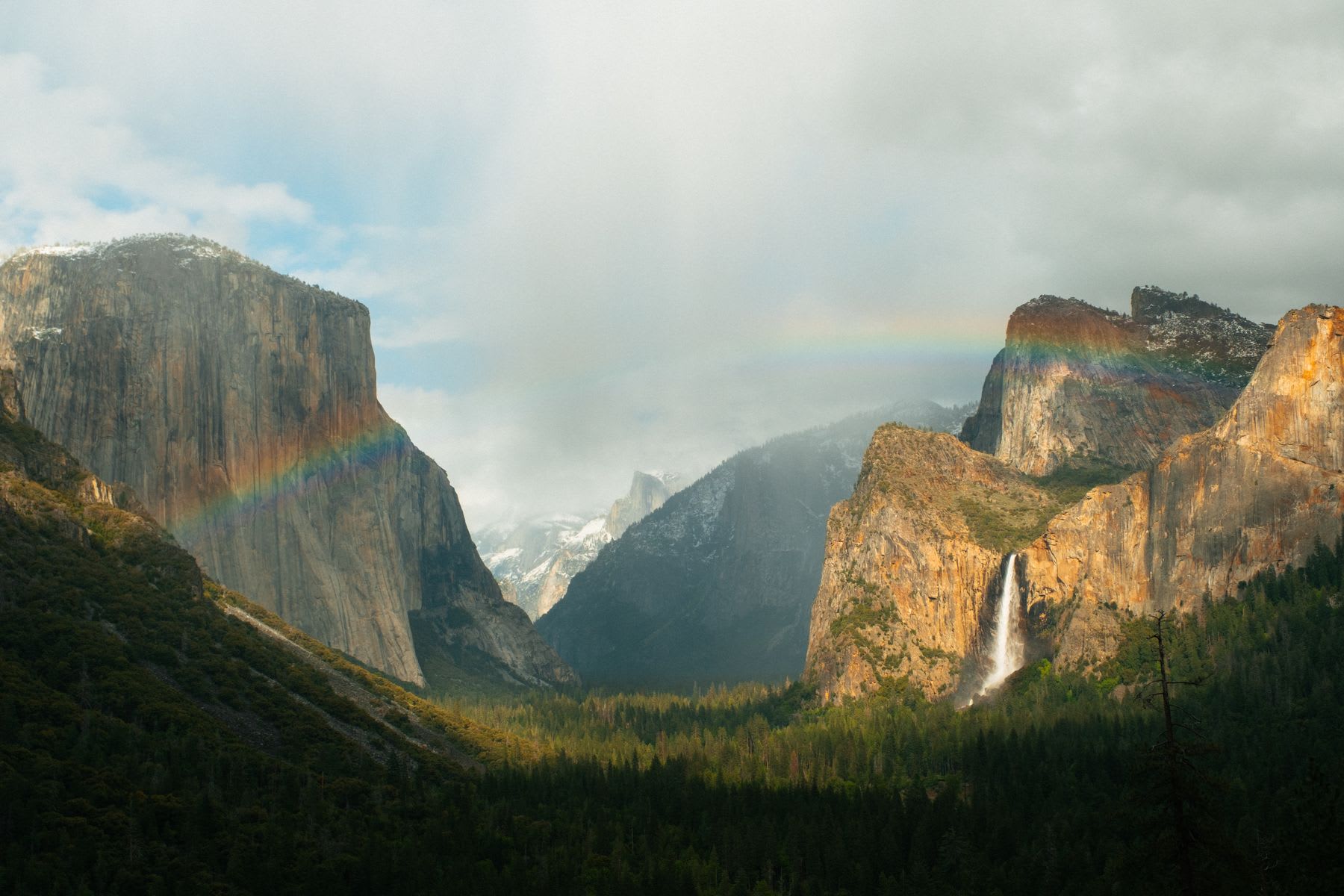 A vivid rainbow stretches across towering granite cliffs and a waterfall in a dramatic forested mountain valley beneath a cloudy sky.