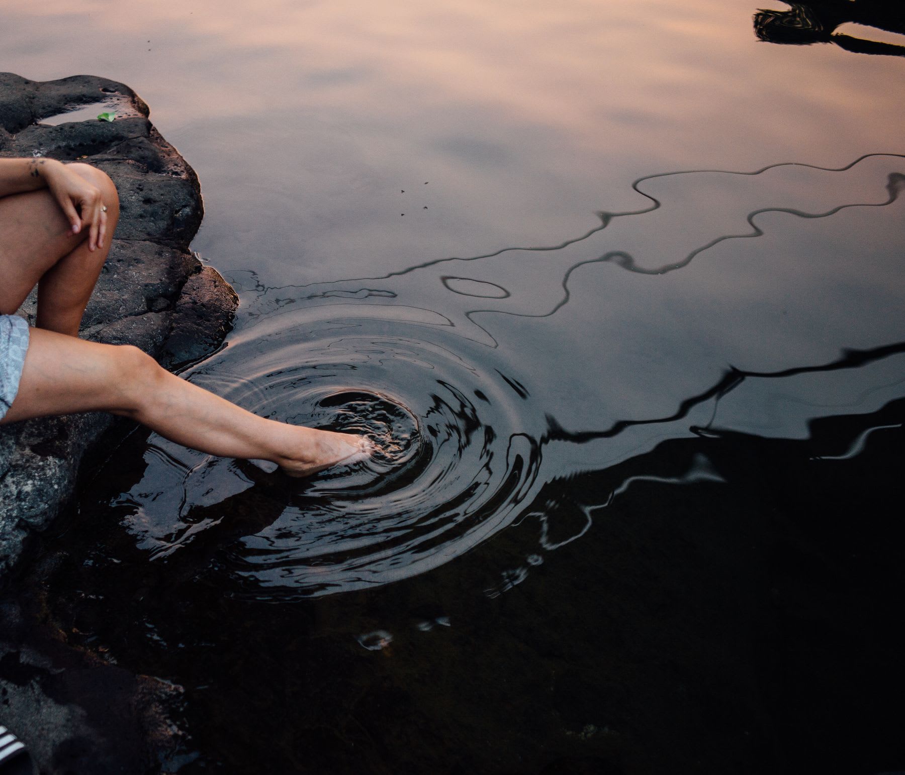 A woman sitting on a rock, with her foot in a body of water.