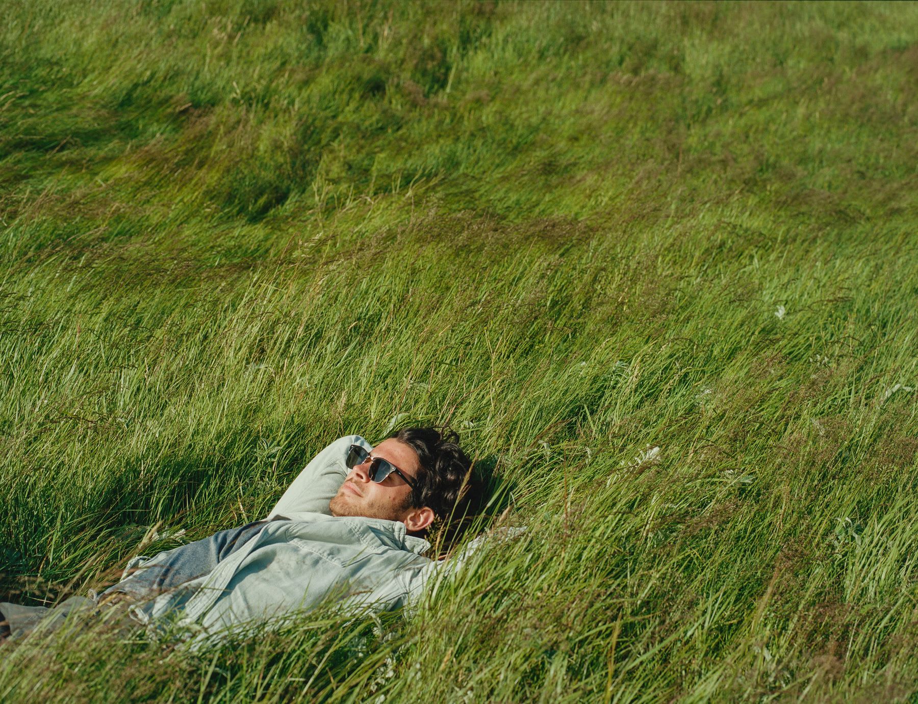 Man in sunglasses and long sleeved shirt lying in a field with tall grass during daytime