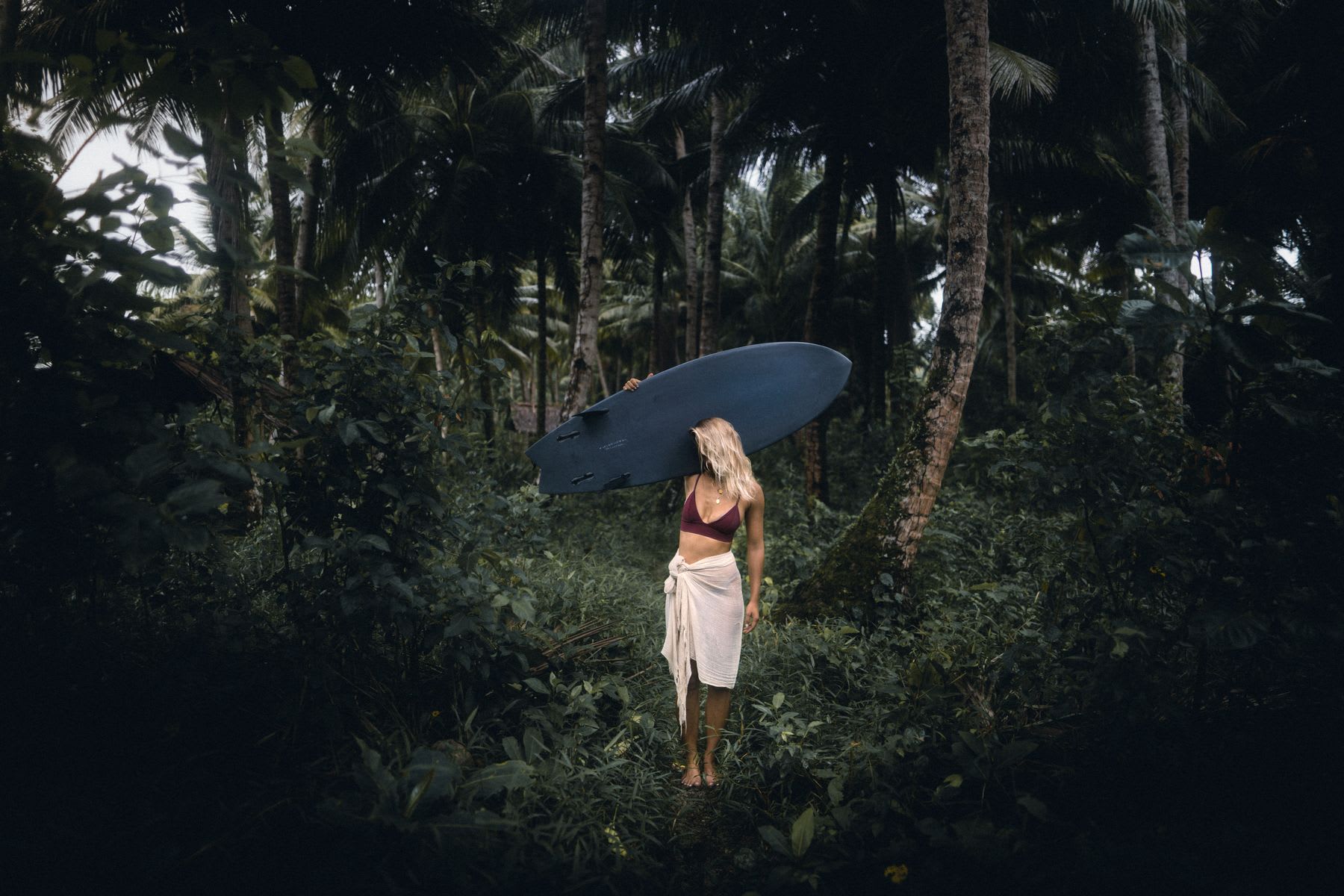 A woman in the middle of a forest holding a blue surfboard and she is looking to the side