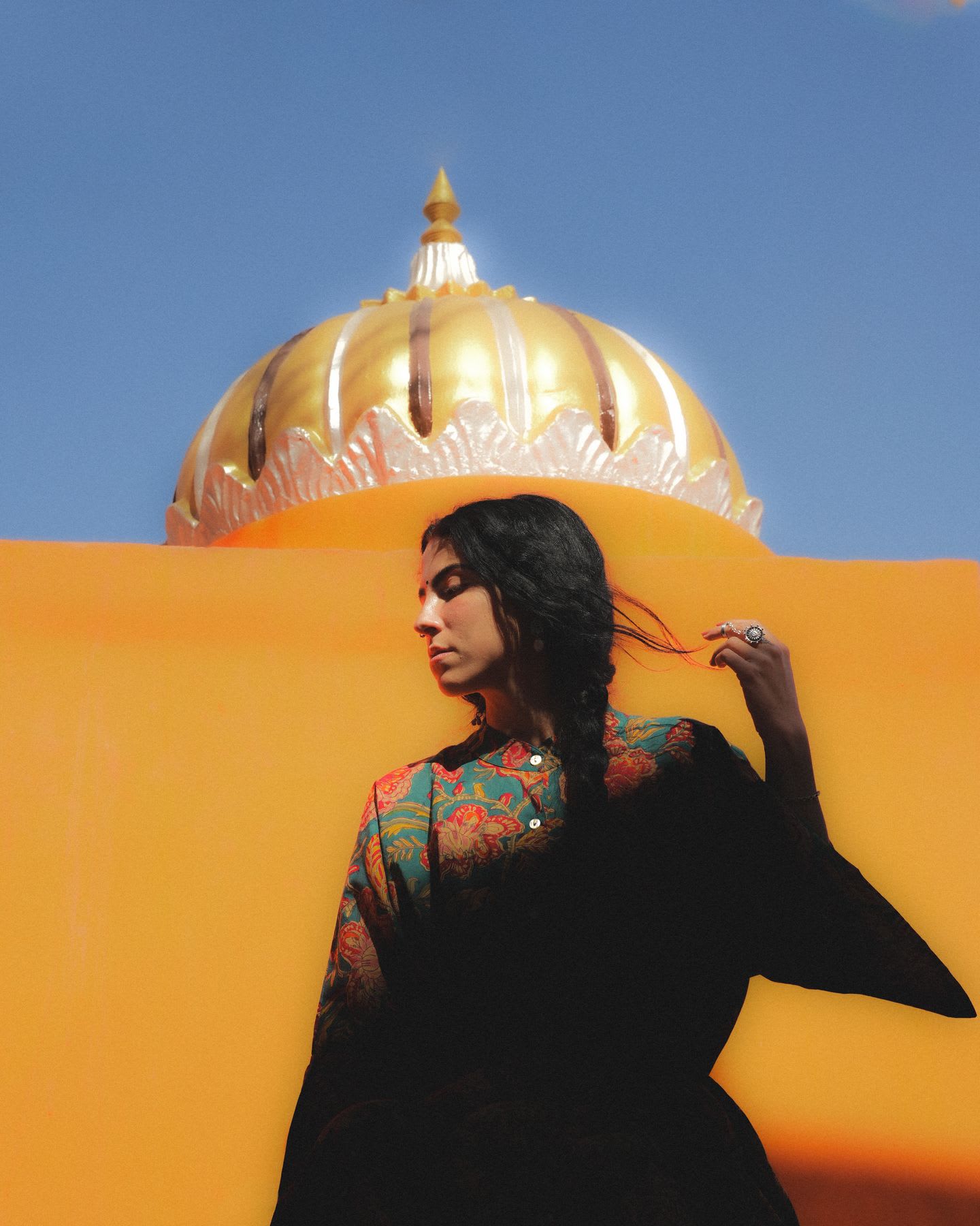 A woman in colorful clothing poses in front of a golden dome and bright orange wall under a clear blue sky.