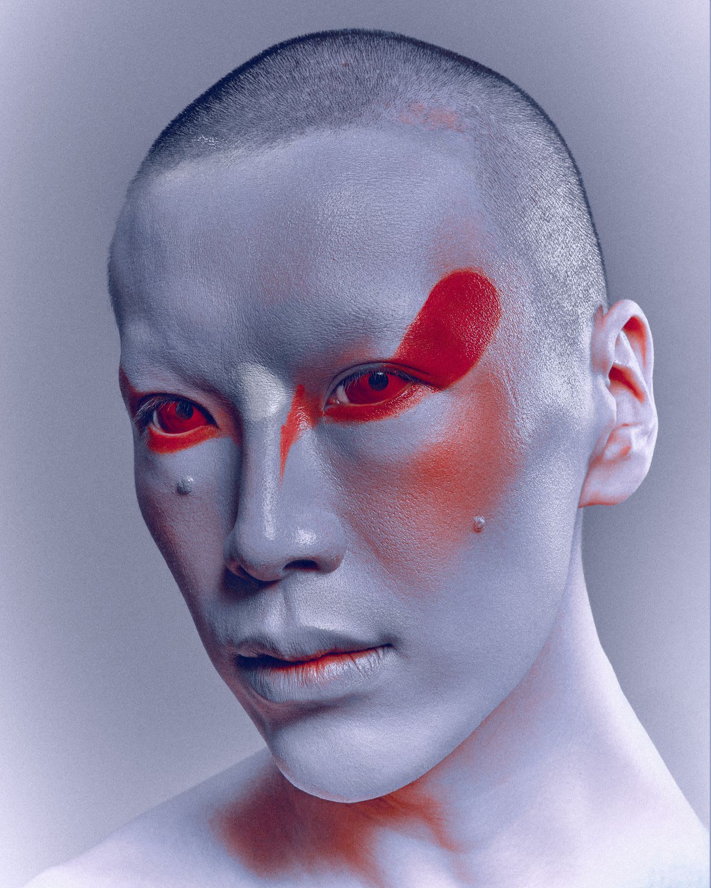 An avant-garde portrait of a person with a shaved head, pale skin, and bold red makeup around their piercing red eyes.
