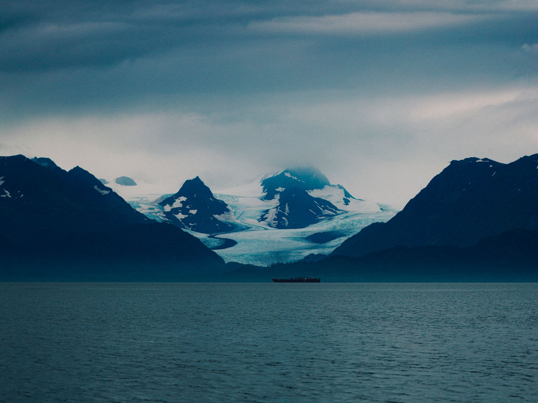 A lone ship sails across a deep blue sea towards a magnificent glacier descending from a snow-covered mountain range.
