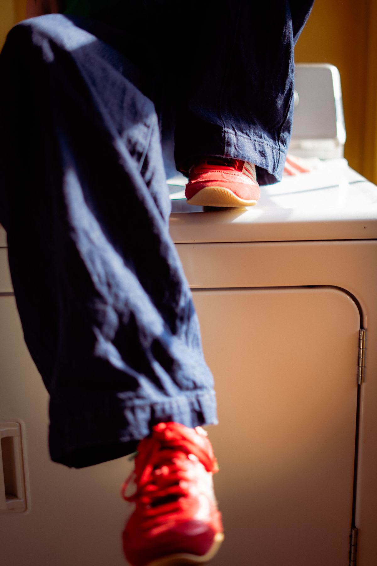 A close-up shot of a person's dangling legs in blue pants and bright red sneakers while sitting on a white appliance.