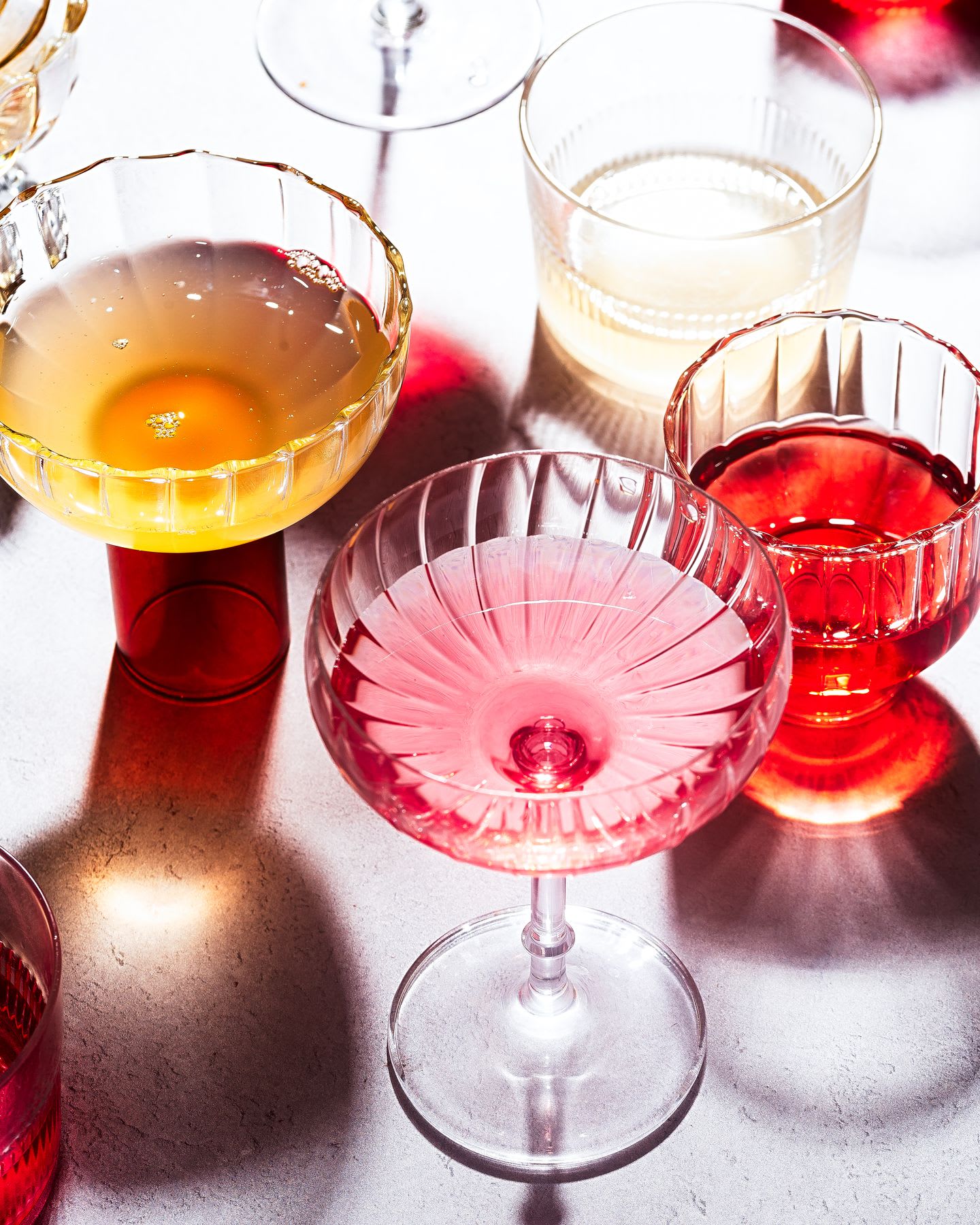 An assortment of colorful cocktails in elegant, patterned glassware casts vibrant shadows on a bright, textured surface.